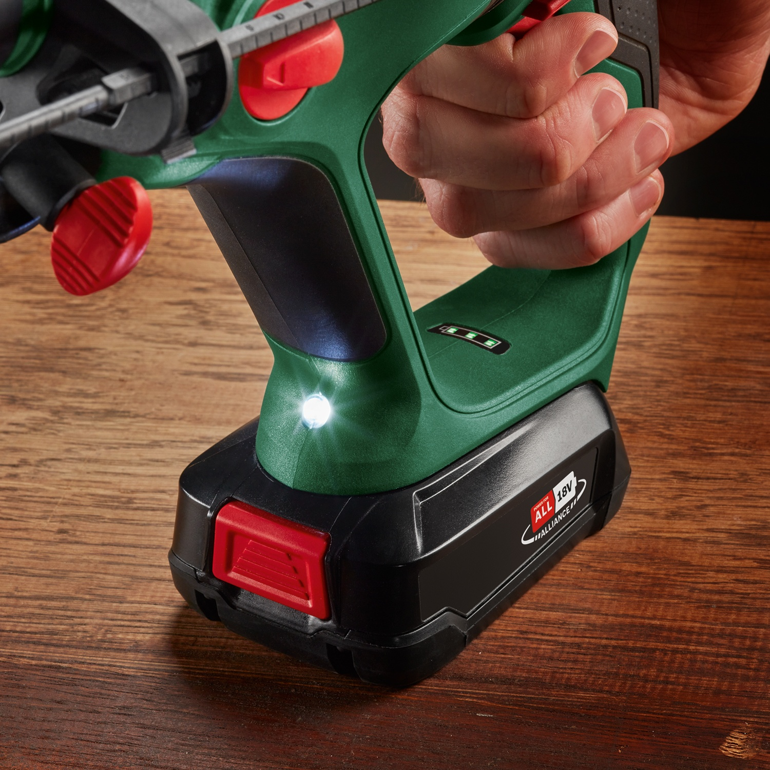 Una mano che tiene un trapano elettrico verde e nero su una superficie di legno. Il trapano ha un pulsante rosso e una luce intensa.