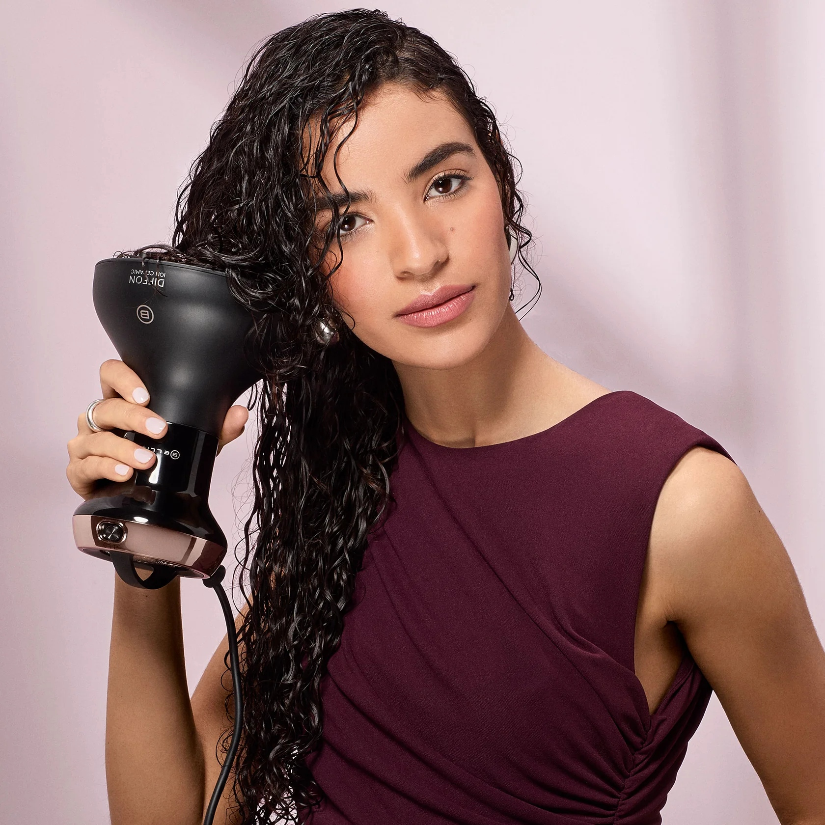 Mujer con cabello rizado usando un secador de pelo negro. Lleva un vestido granate, fondo rosa.