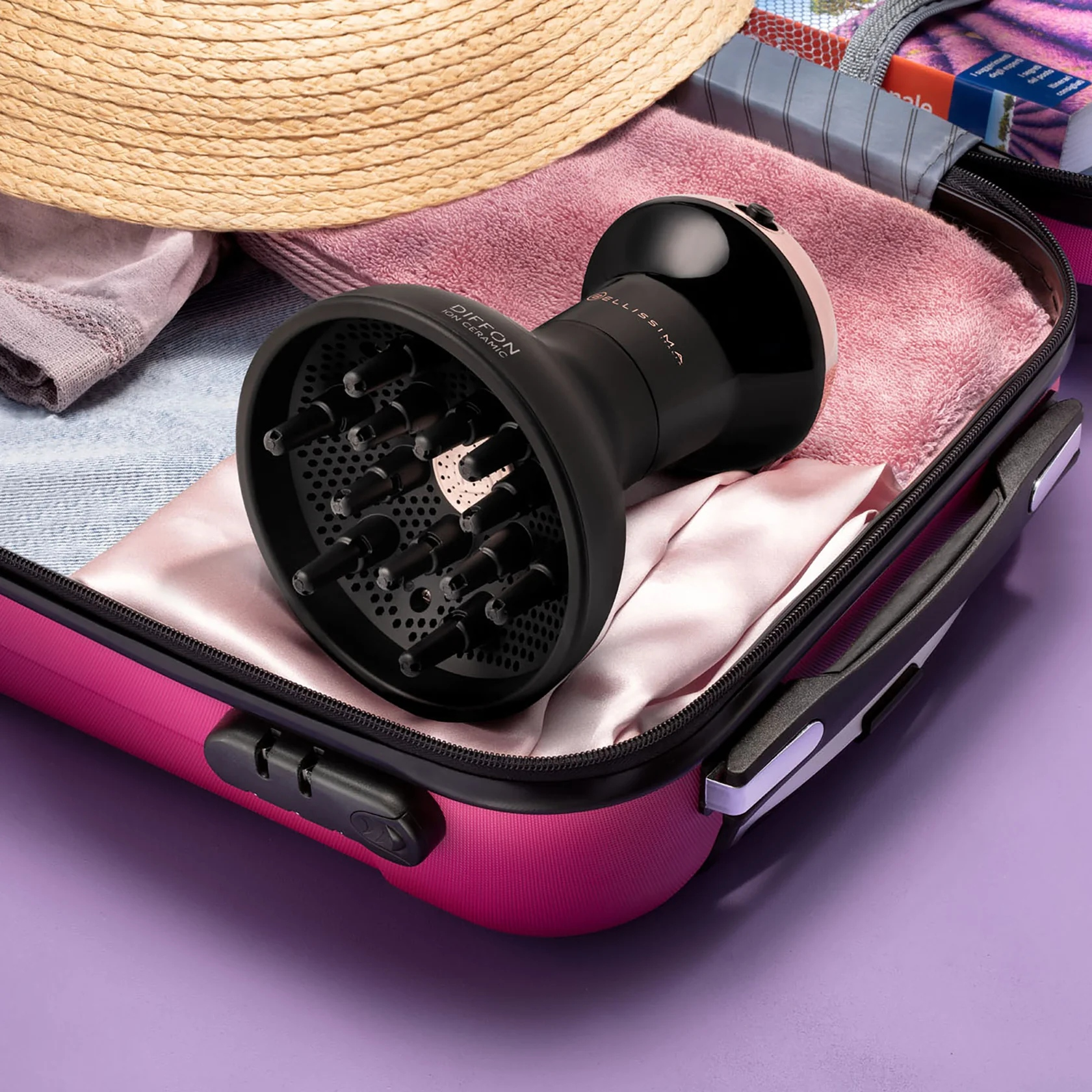Un difusor negro de secador de pelo está en una maleta rosa con un sombrero de paja y una toalla rosa. El fondo es morado.