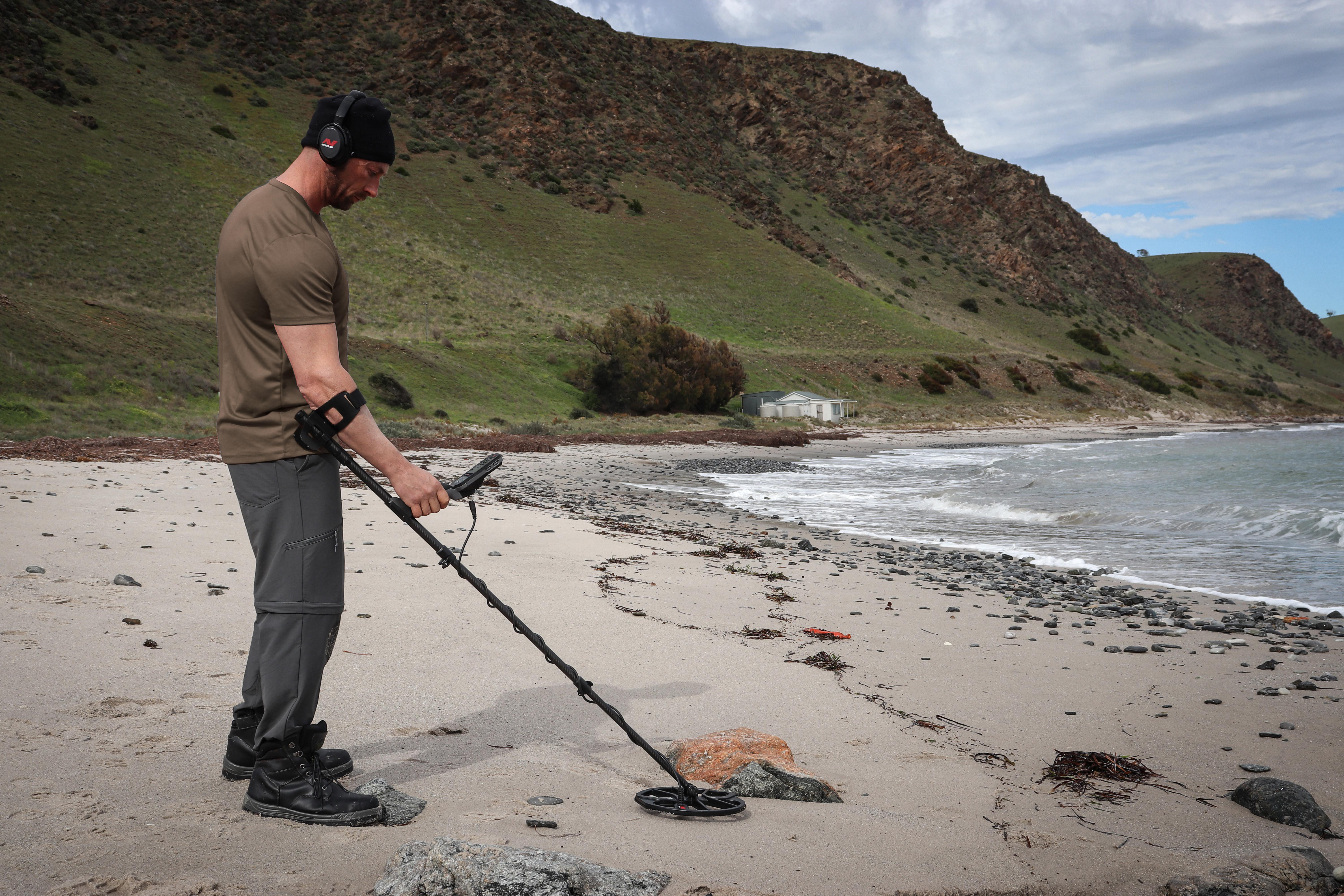 Mann mit Metalldetektor am Strand. Berge und Ozean im Hintergrund. Bewölkter Himmel.