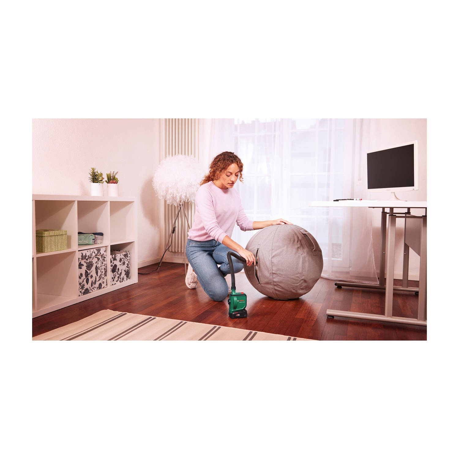 Donna che usa un aspirapolvere verde su un pouf grigio in una stanza con scrivania e scaffali bianchi. Il pavimento è in legno.