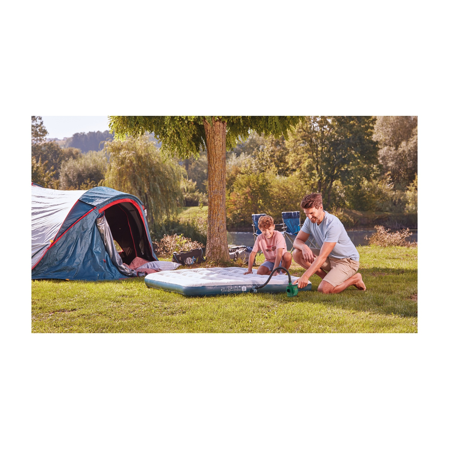 Un uomo e un ragazzo gonfiano un materasso ad aria vicino a una tenda. La scena è un campo erboso con alberi e un fiume sullo sfondo.
