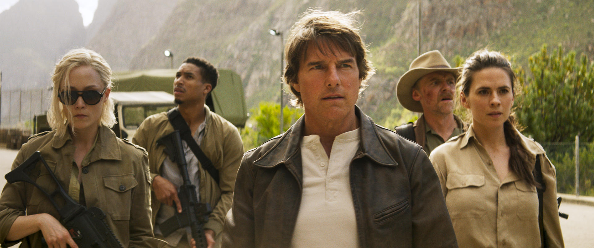 Eine Gruppe von Personen, darunter Tom Cruise, geht draußen. Einige halten Waffen. Berge und Grün sind im Hintergrund.
