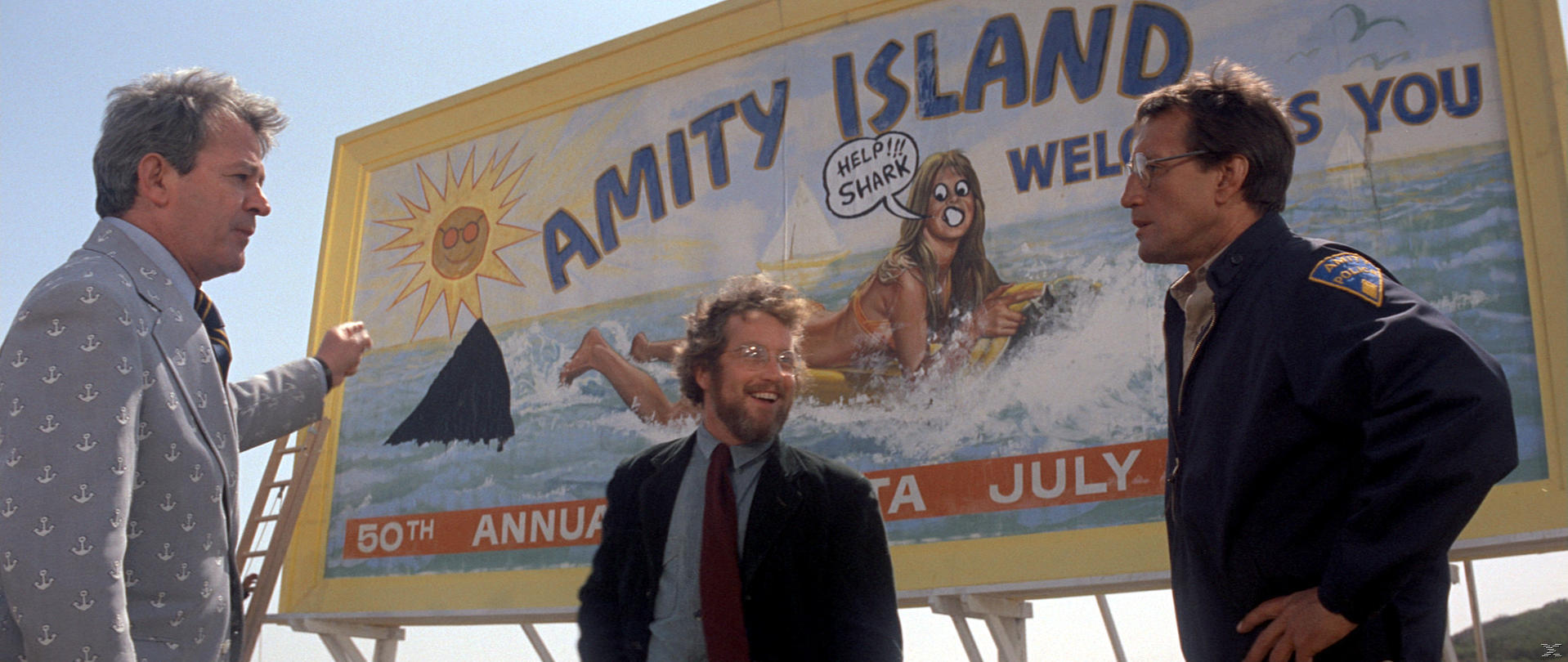 Drei Männer stehen in der Nähe einer Werbetafel von 'Amity Island' mit einem Hai-Bild. Das Schild sagt: 'Hilfe!!! Hai.' und '50. Jahrestag.'