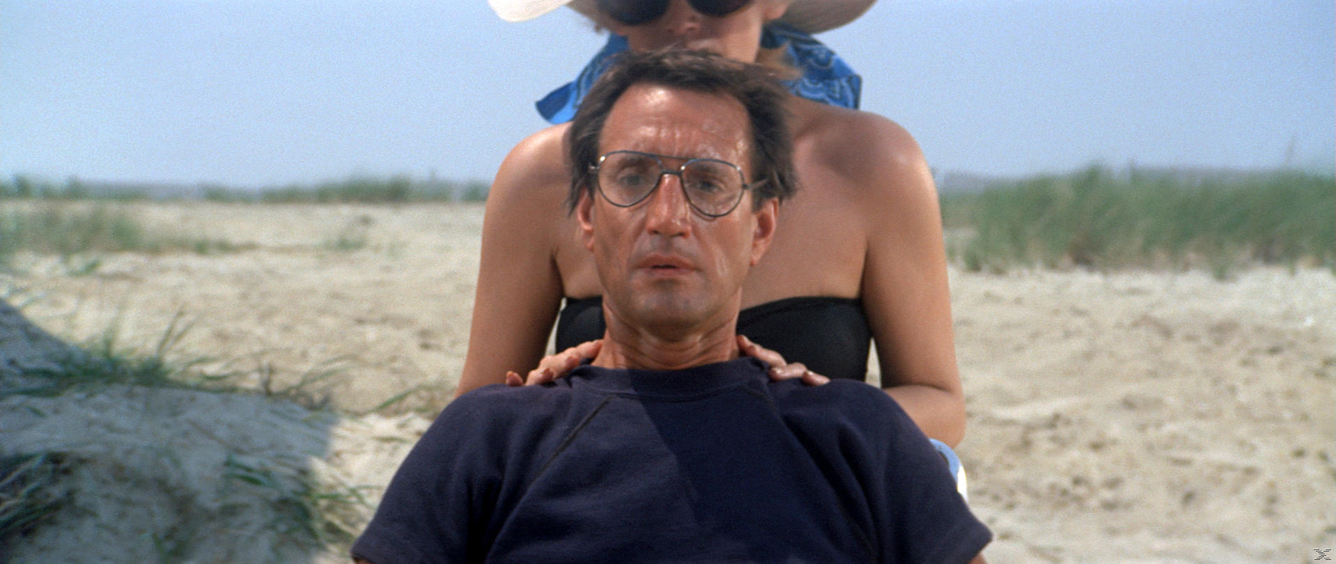 Mann mit Brille sitzt am Strand, Frau hinter ihm. Klarer Himmel, Dünen und Sand im Hintergrund.