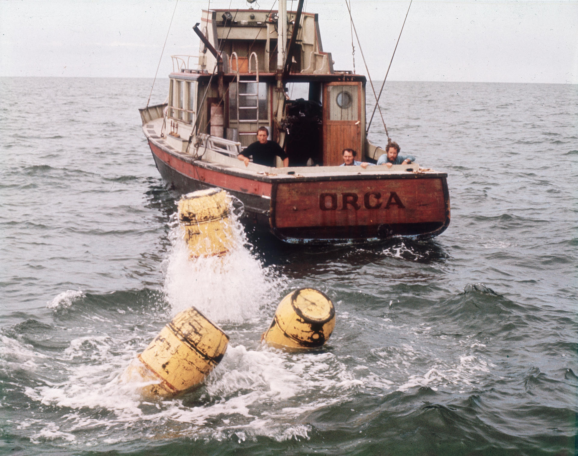 Ein Boot namens Orca mit drei Männern an Bord, umgeben von Wasser. Gelbe Fässer schwimmen in der Nähe, mit Spritzern.