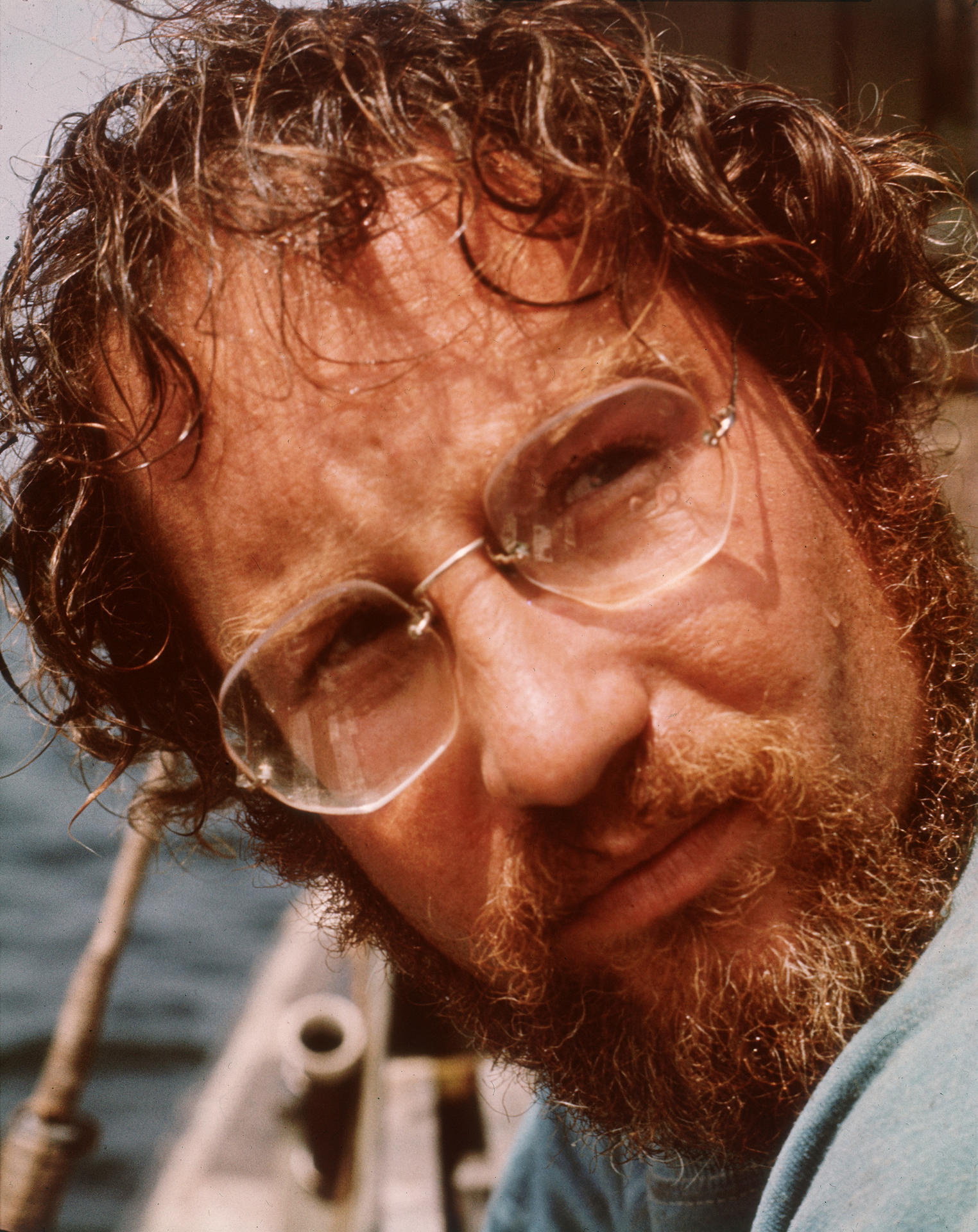 Nahaufnahme eines Mannes mit rotem Haar, Bart und Brille. Er blickt nach unten, anscheinend auf einem Boot mit blauem Hintergrund.