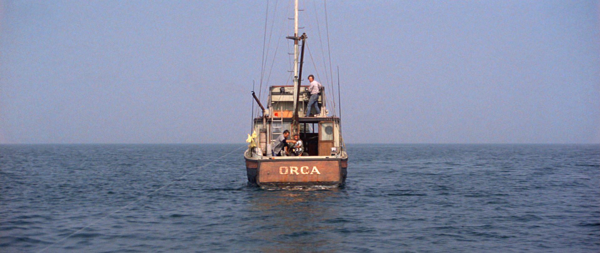 Das Fischerboot Orca fährt auf dem Meer. Männer sind auf dem Boot. Der Himmel ist klar. Das Boot ist braun mit dem Wort ORCA darauf.