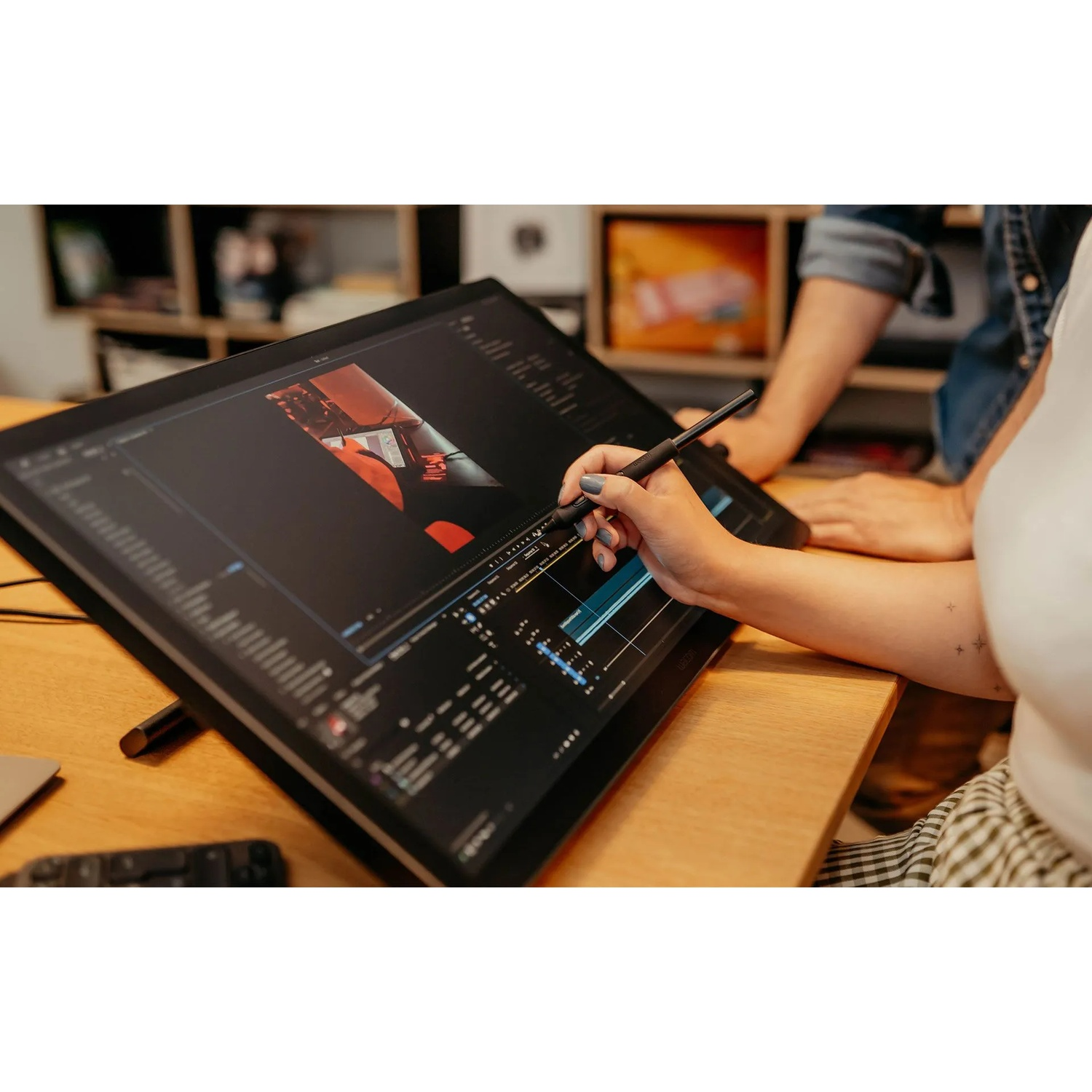 Una persona usa una penna su un grande schermo che mostra un software di editing video. Un'altra persona è accanto a loro. Tavolo di legno.