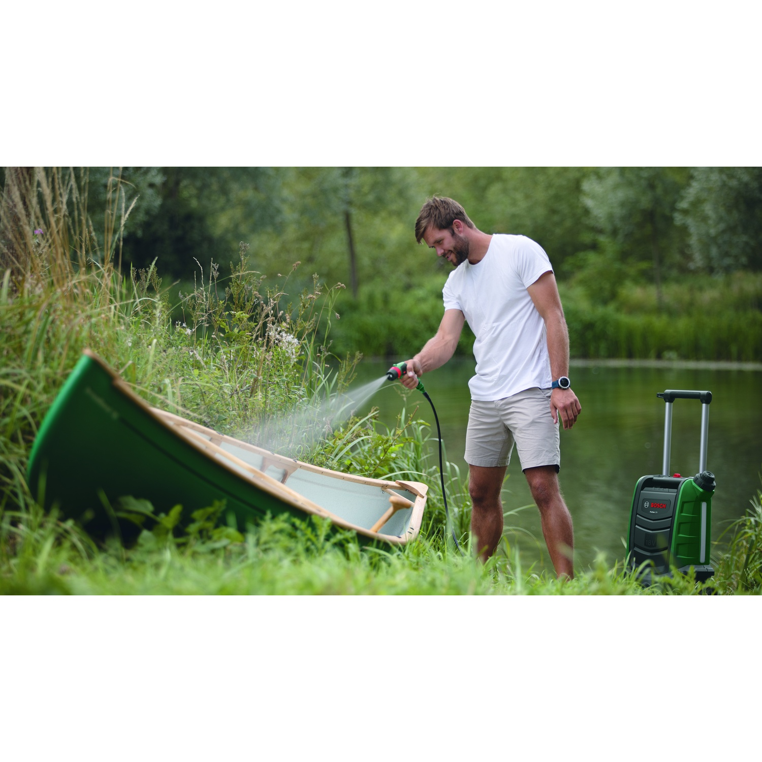 Uomo che lava una canoa verde vicino a un lago, usando un'idropulitrice. Una vegetazione lussureggiante lo circonda. Un'idropulitrice è a destra.