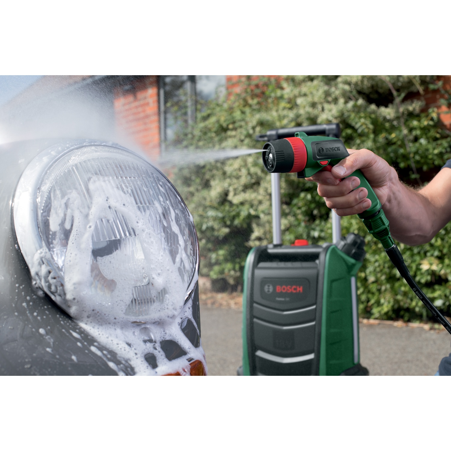 Una persona lava un faro di un'auto con un'idropulitrice Bosch. L'auto è grigia, l'idropulitrice verde e nera. Sapone schiumoso ricopre la luce.
