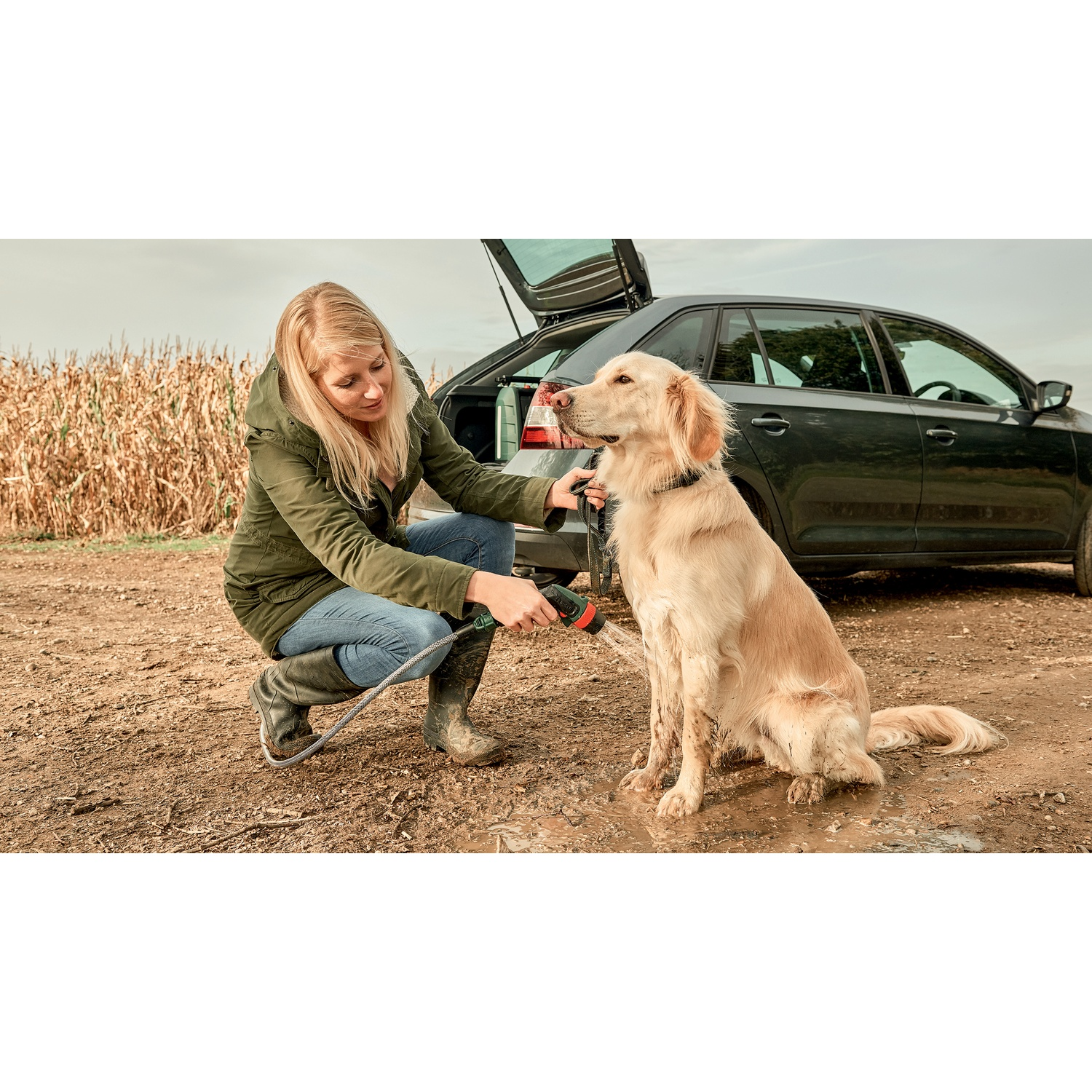 Una donna lava un golden retriever con un tubo. Il cane è seduto vicino a un'auto con il bagagliaio aperto su un terreno fangoso.