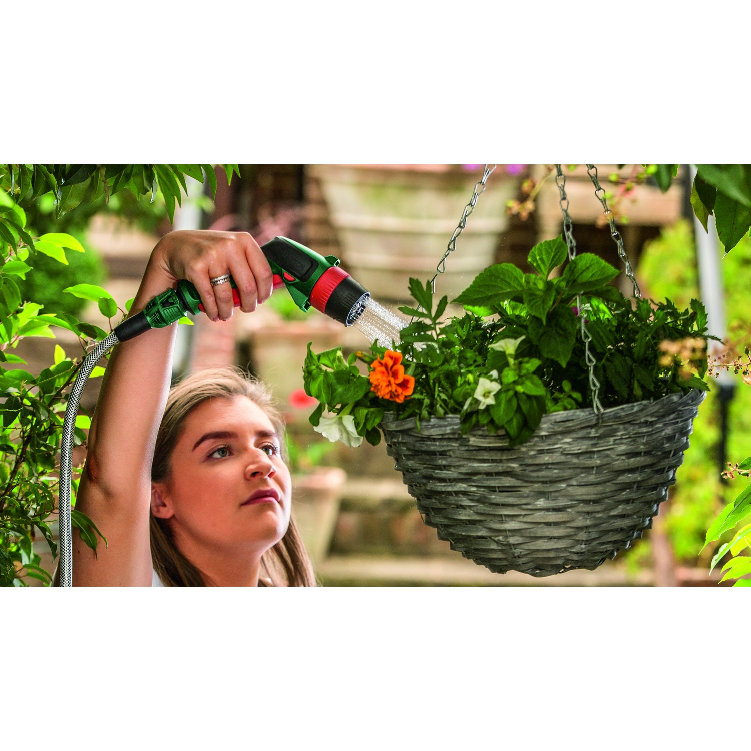 Una donna innaffia un cestino appeso con un tubo. Il cestino contiene fiori. Lo sfondo include vegetazione e una struttura in mattoni.