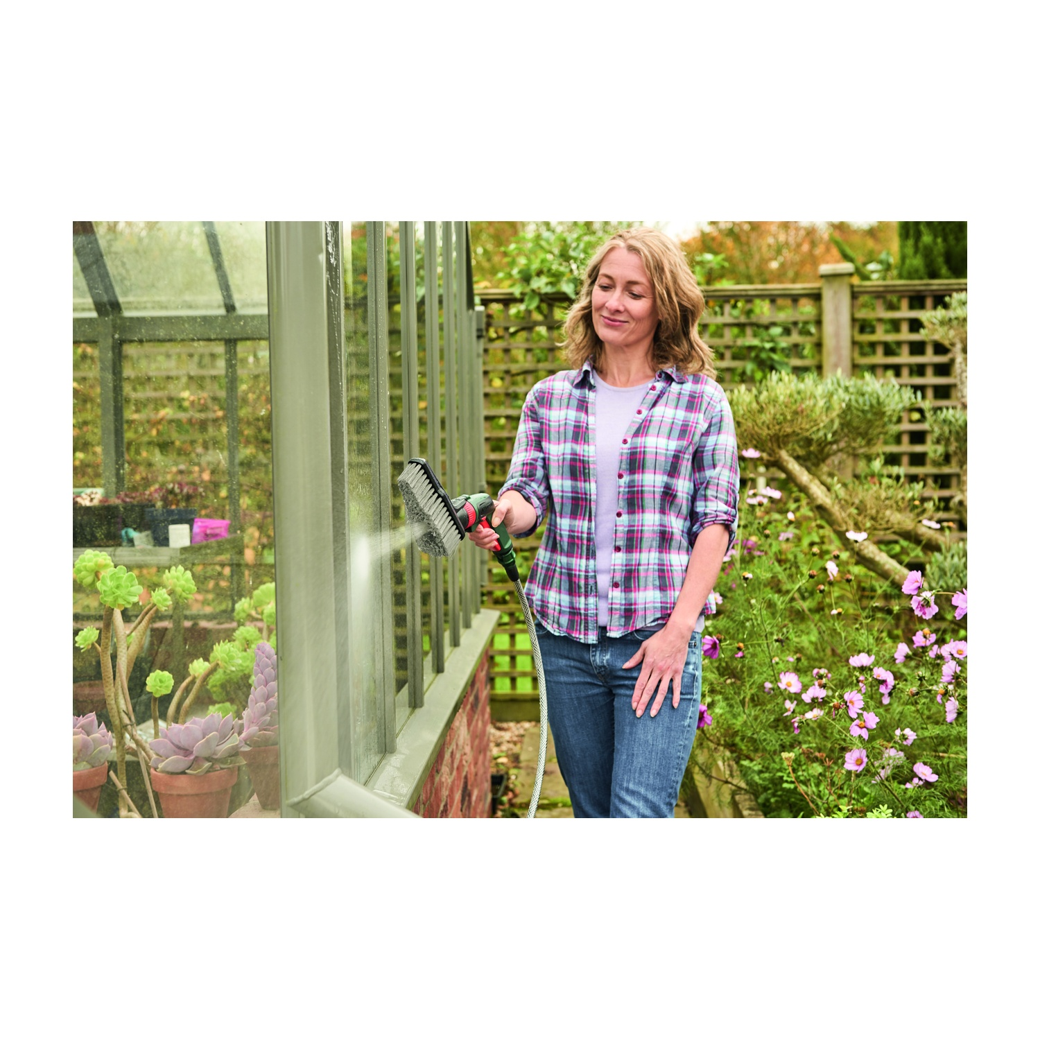 Una donna con una camicia a quadri spruzza acqua su una serra. Piante sullo sfondo. Sorride mentre fa giardinaggio.