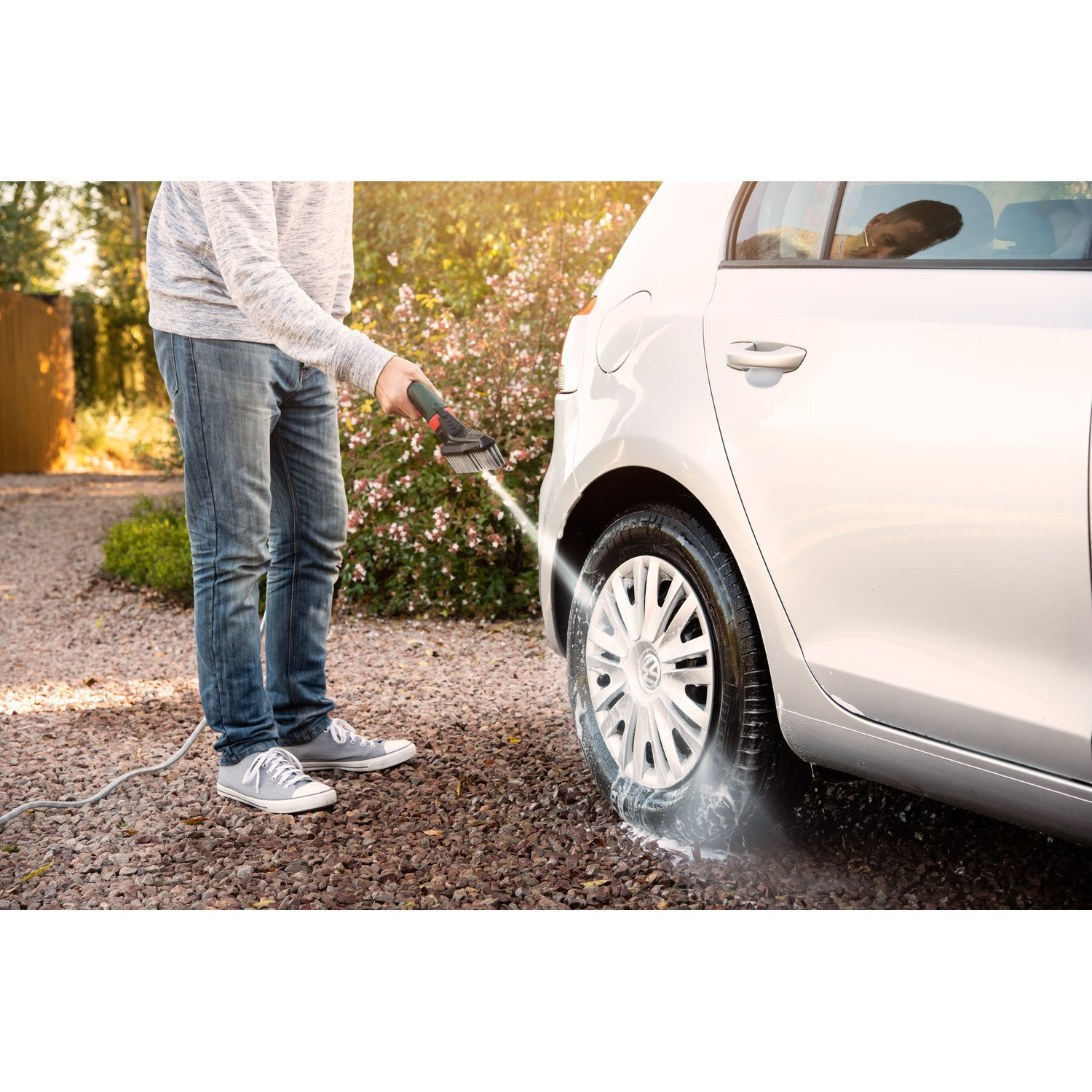 Una persona lava un'auto argentata con un'idropulitrice. L'acqua schizza sullo pneumatico. Lo sfondo mostra il fogliame e la luce del sole.