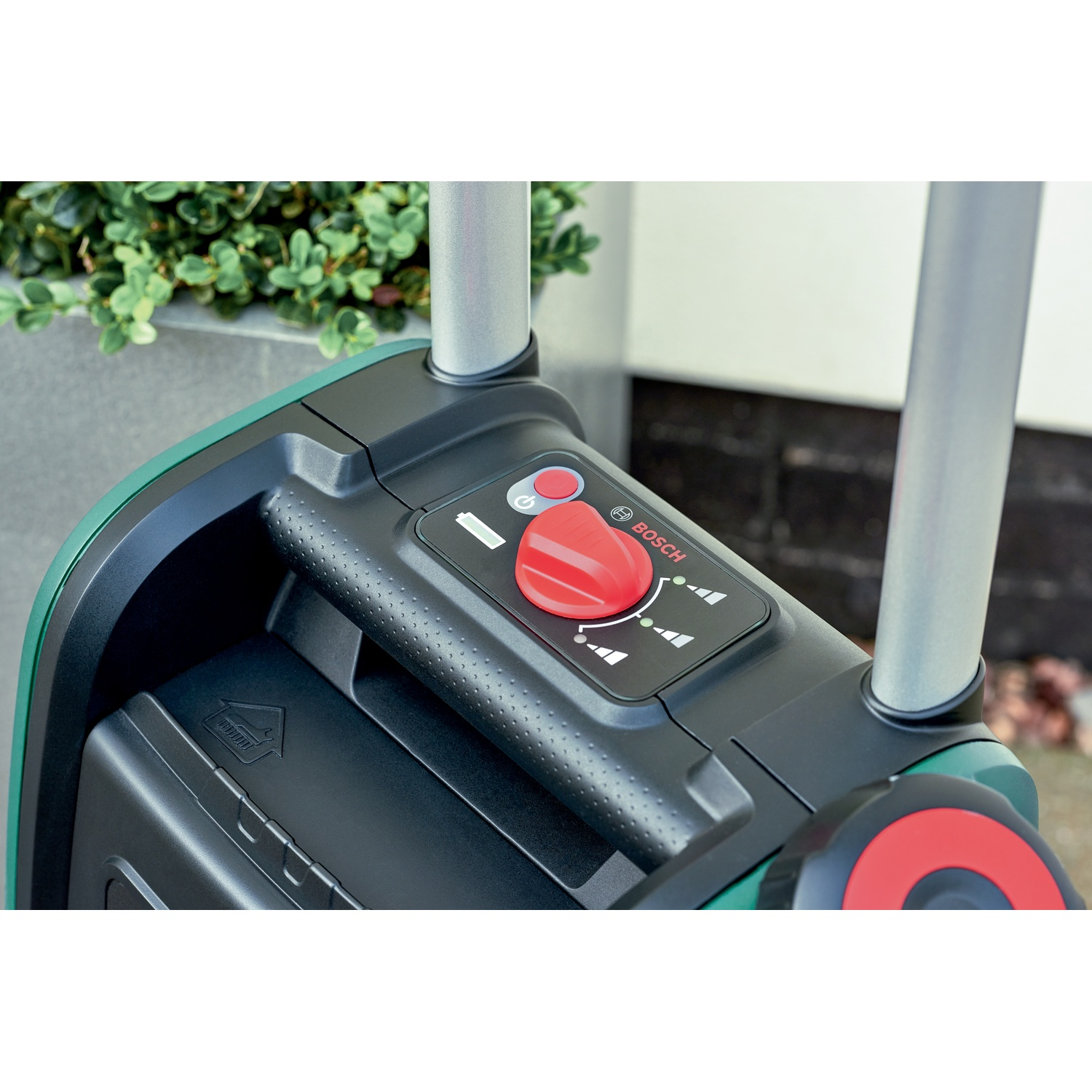 Primo piano di uno strumento Bosch nero e verde con un quadrante rosso. Manici argentati e una pianta verde sullo sfondo.