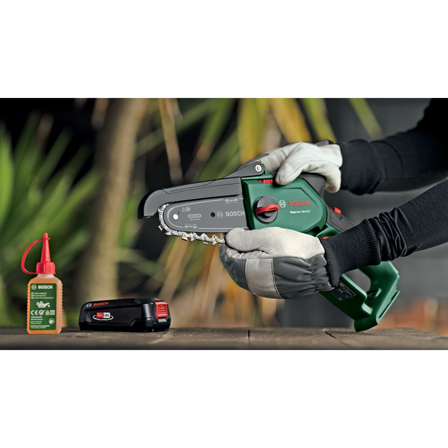Una persona tiene in mano una motosega Bosch verde e nera. Olio e batteria su un tavolo di legno. Lo sfondo ha un fogliame verde.