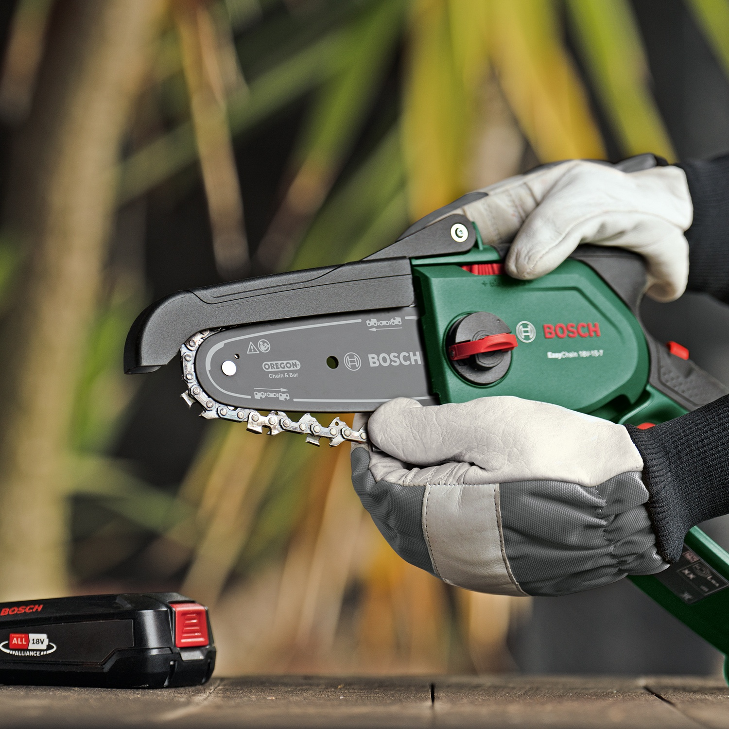 Una persona tiene in mano una motosega Bosch verde e nera con guanti bianchi. Una batteria è su una superficie di legno.