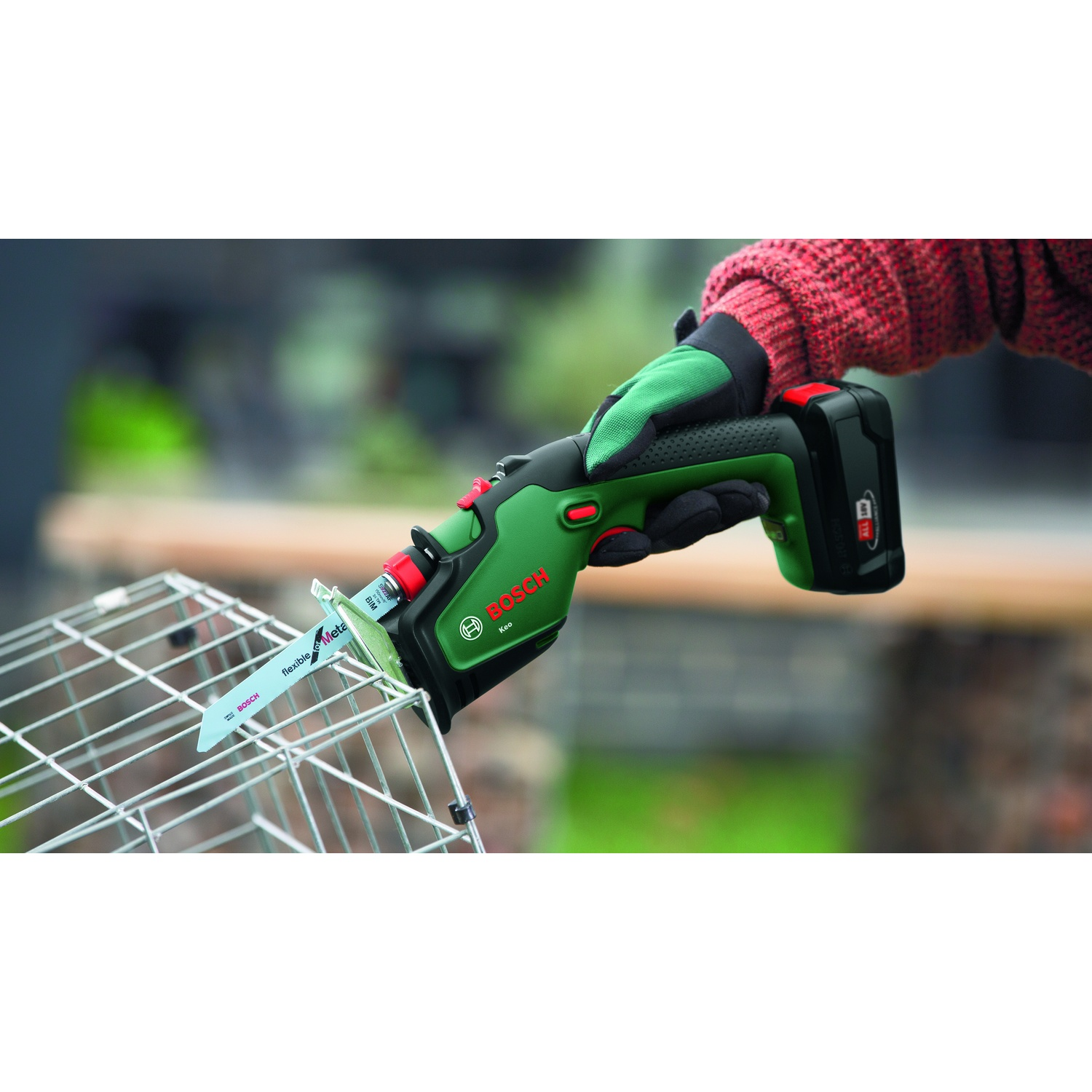 Una persona tiene in mano un seghetto Bosch verde e nero che taglia una gabbia di metallo. La mano indossa un guanto verde. È visibile la manica rossa del maglione.
