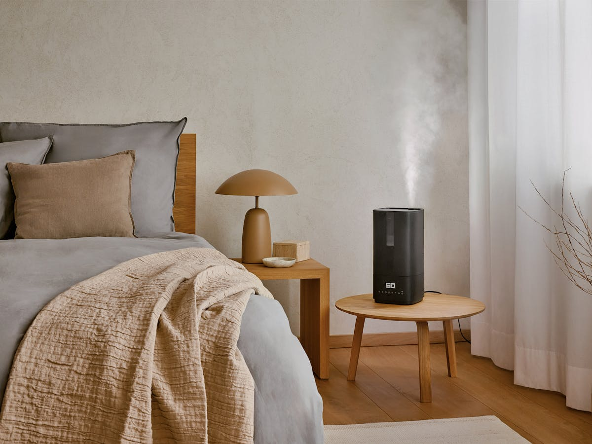 Scena di camera da letto con letto, tavolo, lampada e umidificatore. Colori neutri e atmosfera accogliente.