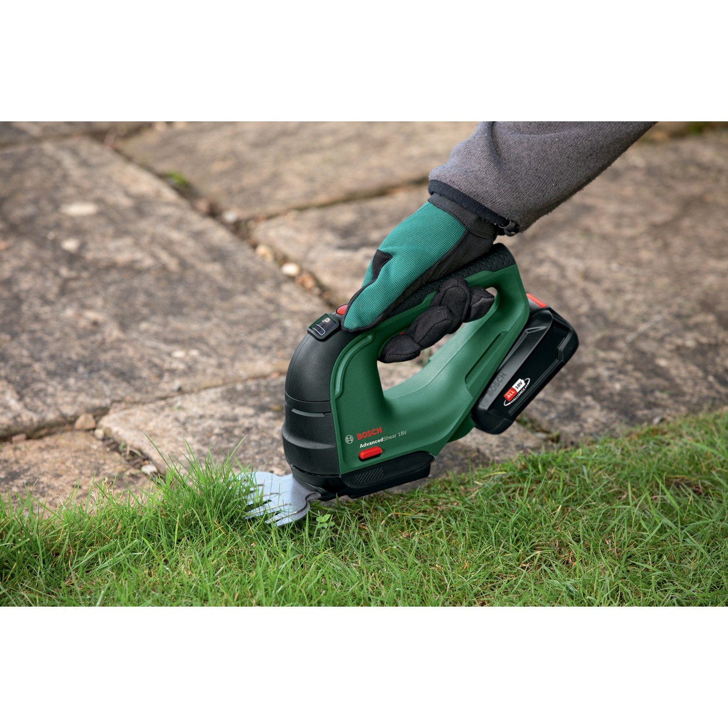 Una mano con un guanto verde tiene un tagliasiepi verde e nero. Il trimmer taglia l'erba su una superficie di pietra.