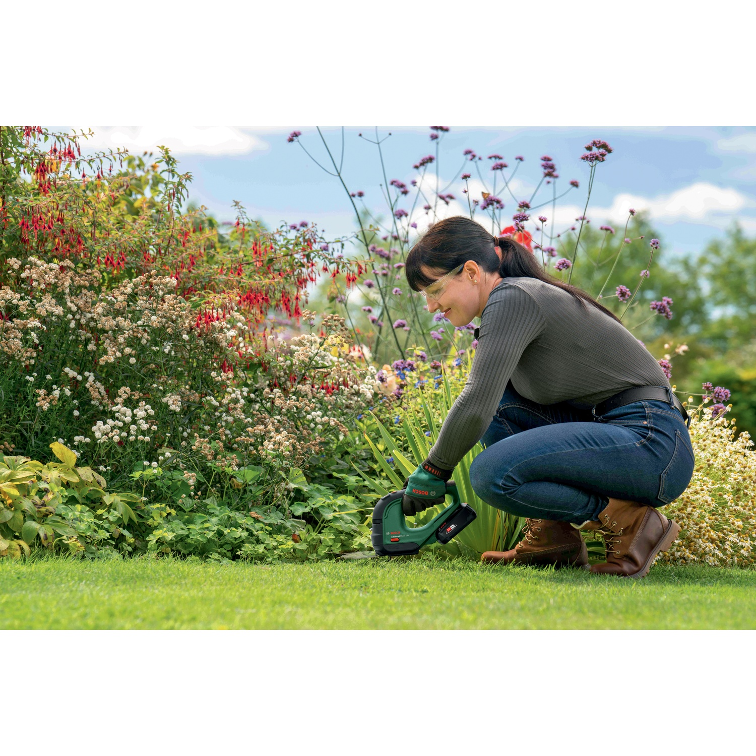 Una donna in giardino taglia l'erba con uno strumento verde. Fiori e piante verdi la circondano in una giornata di sole.