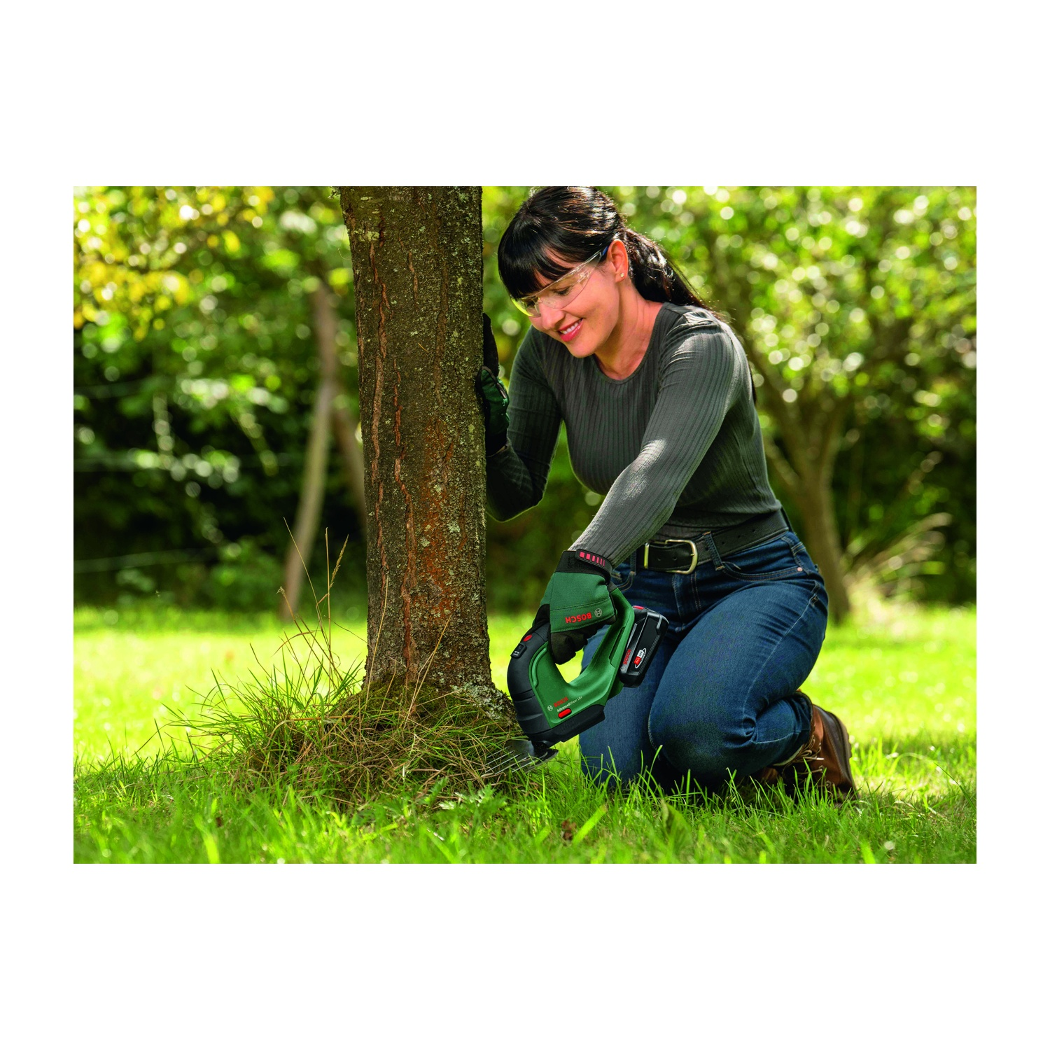 Una donna è in ginocchio, usa una sega verde su un tronco d'albero in un'area erbosa. Indossa occhiali protettivi, guanti e jeans.