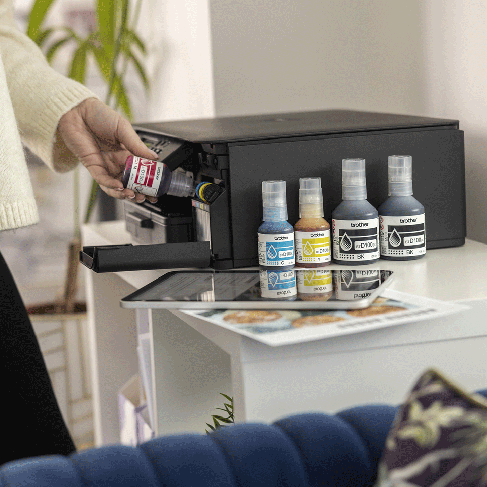 Osoba uzupełnia drukarkę butelkami z tuszem. Cztery butelki z tuszem stoją na białym stole. Obecny jest tablet i wydruki.