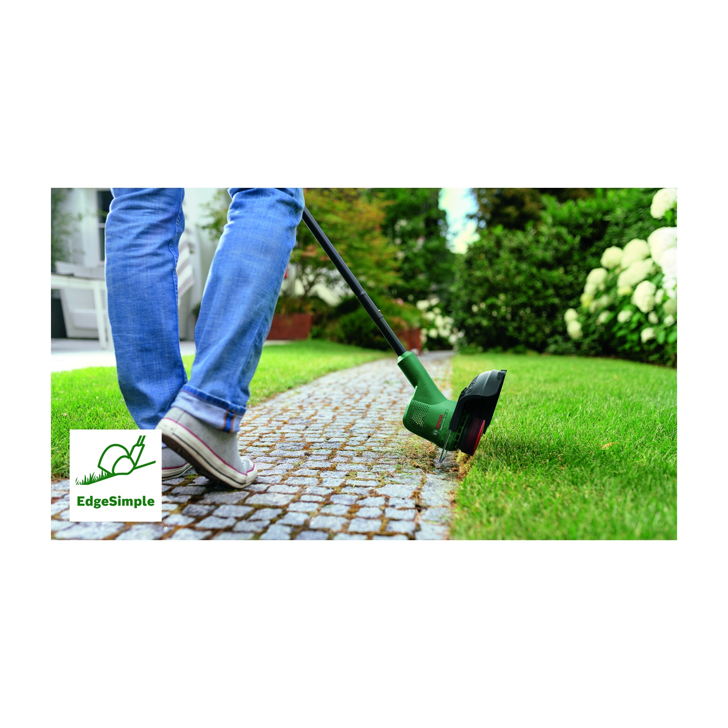 Una persona usa un tagliaerba sul prato. La persona indossa jeans e scarpe da ginnastica. Erba verde e un sentiero lastricato sono visibili.