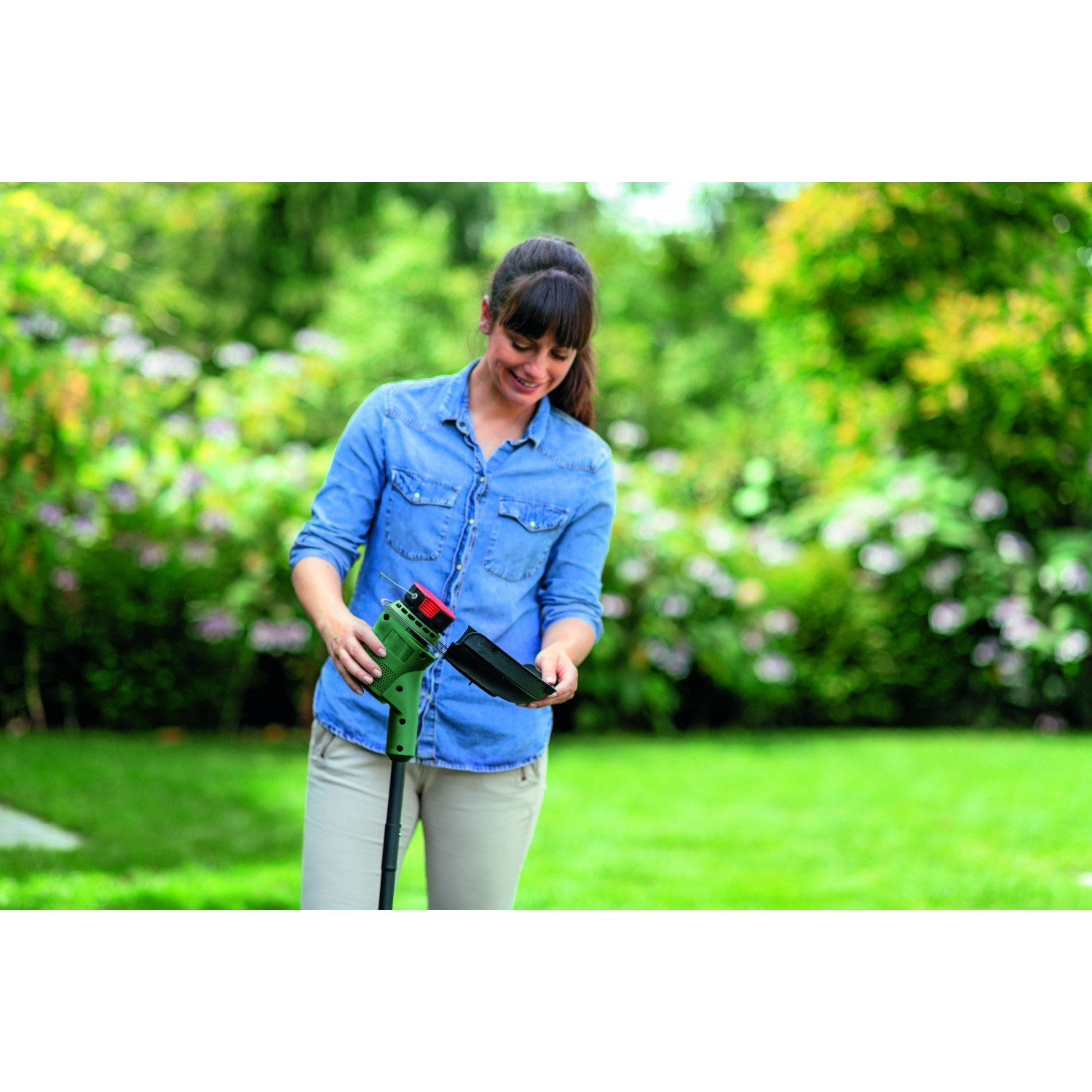 Una donna con una camicia blu e pantaloni beige tiene in mano un attrezzo da giardinaggio verde in un giardino.
