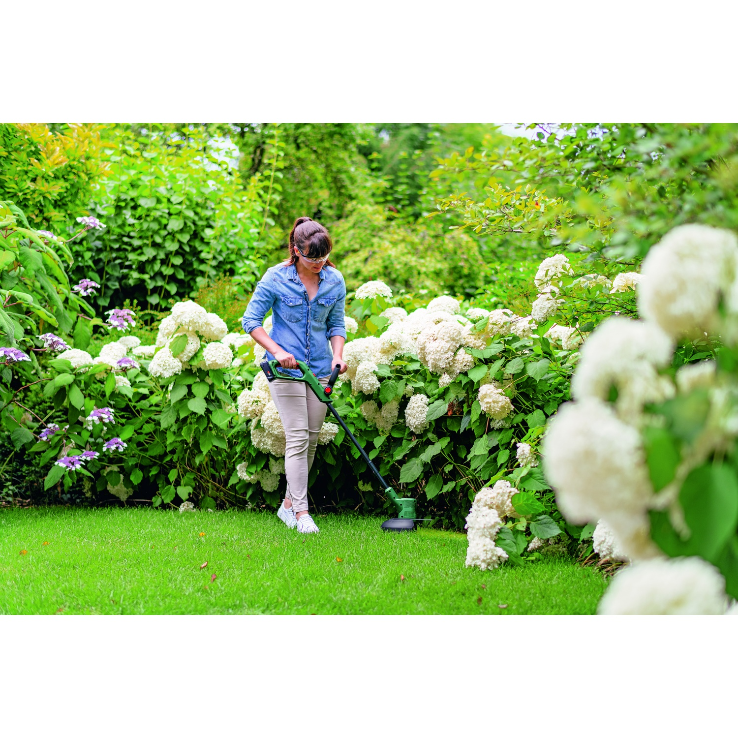 Una donna in giardino taglia l'erba con un tagliaerba verde e nero. Fiori bianchi e cespugli verdi la circondano in una giornata di sole.