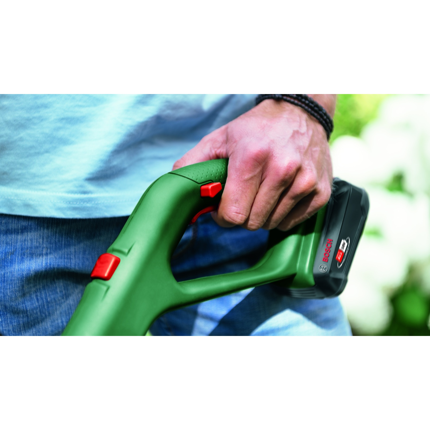 Una persona tiene in mano un attrezzo da giardinaggio Bosch verde con un pulsante rosso. È collegata una batteria nera. Lo sfondo è verde sfocato.