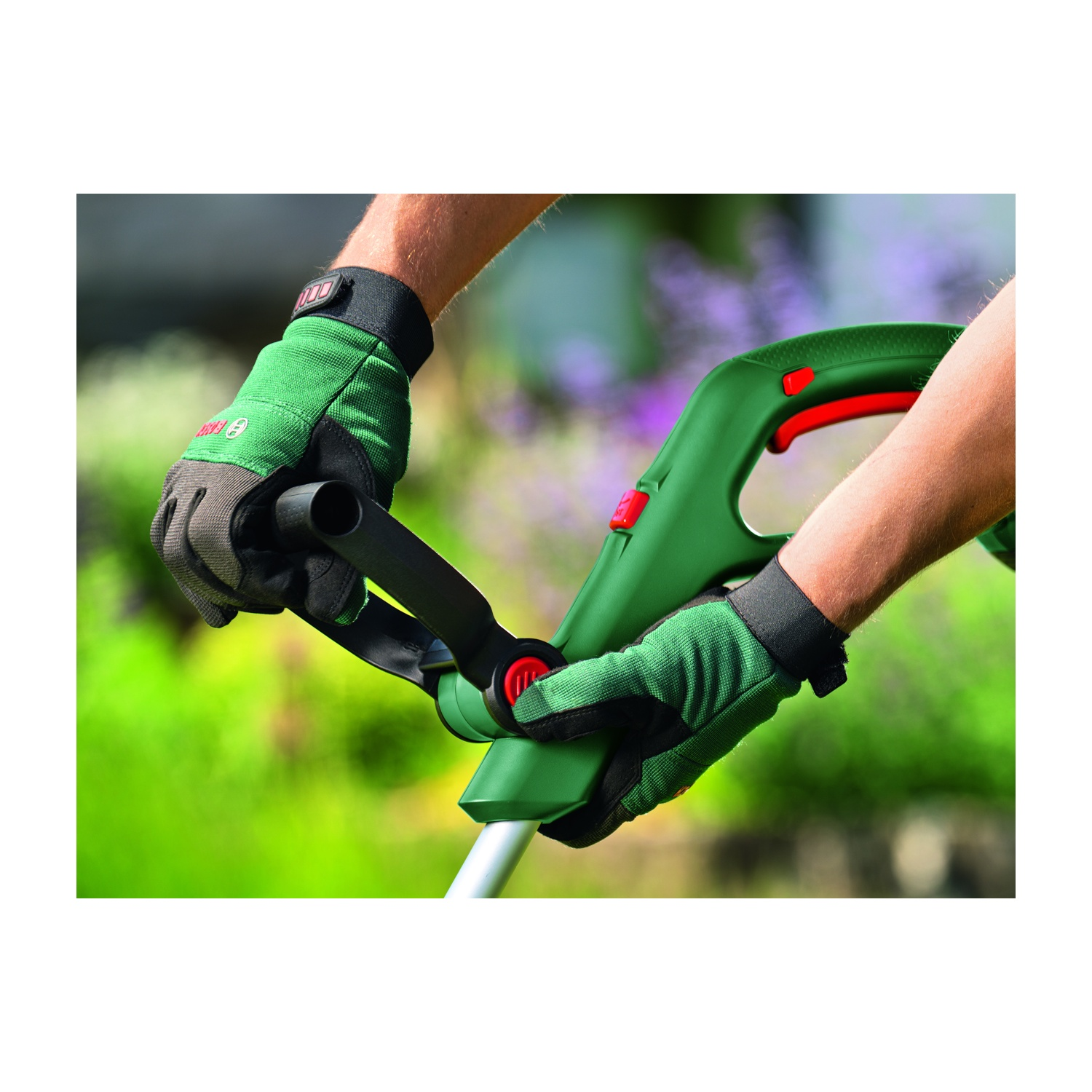 Mani con guanti verdi che tengono un attrezzo da giardinaggio verde e nero. Lo sfondo mostra piante verdi e viola sfocate.