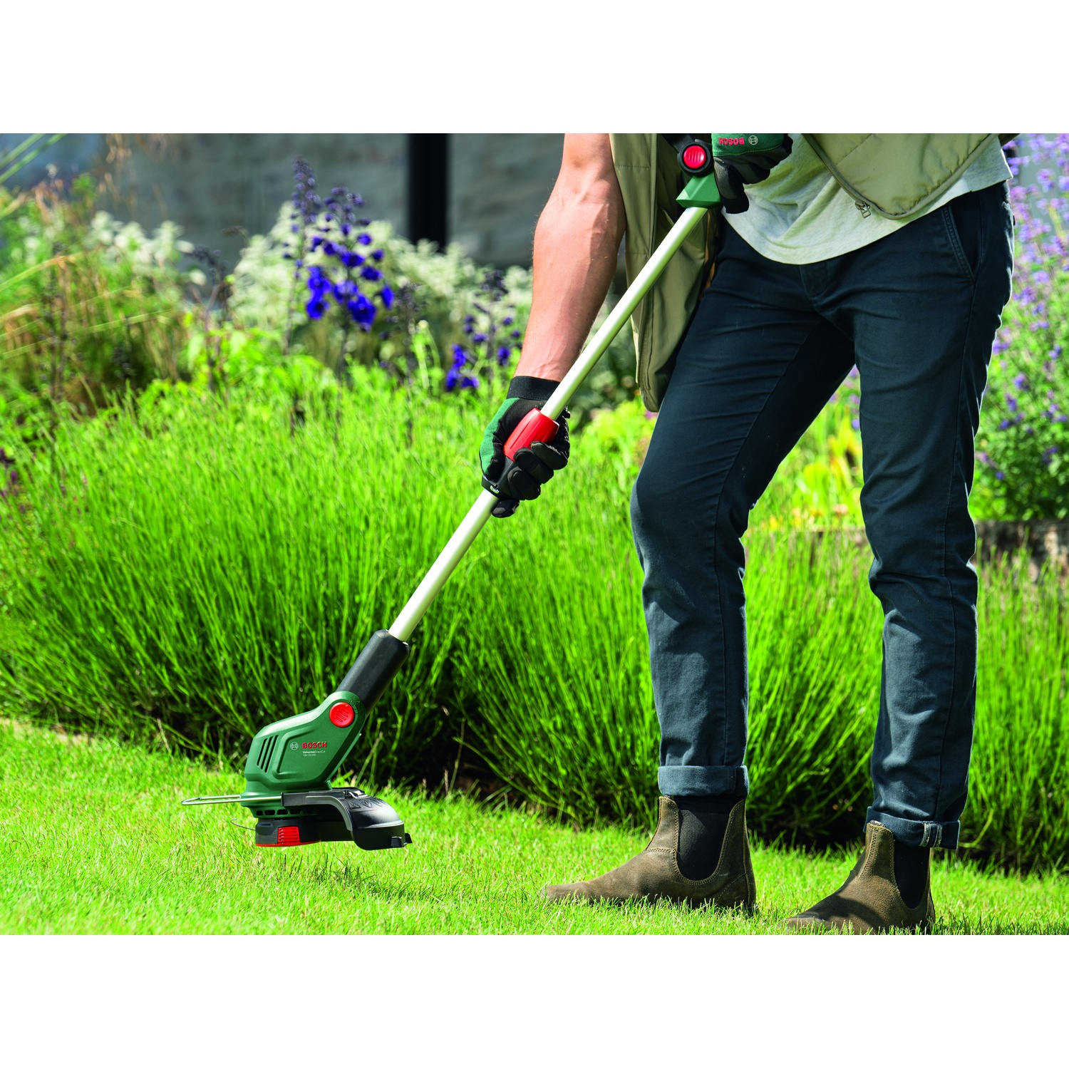 Una persona sta usando un decespugliatore verde e nero. La persona è in piedi sull'erba verde, circondata da arbusti verdi e fiori.