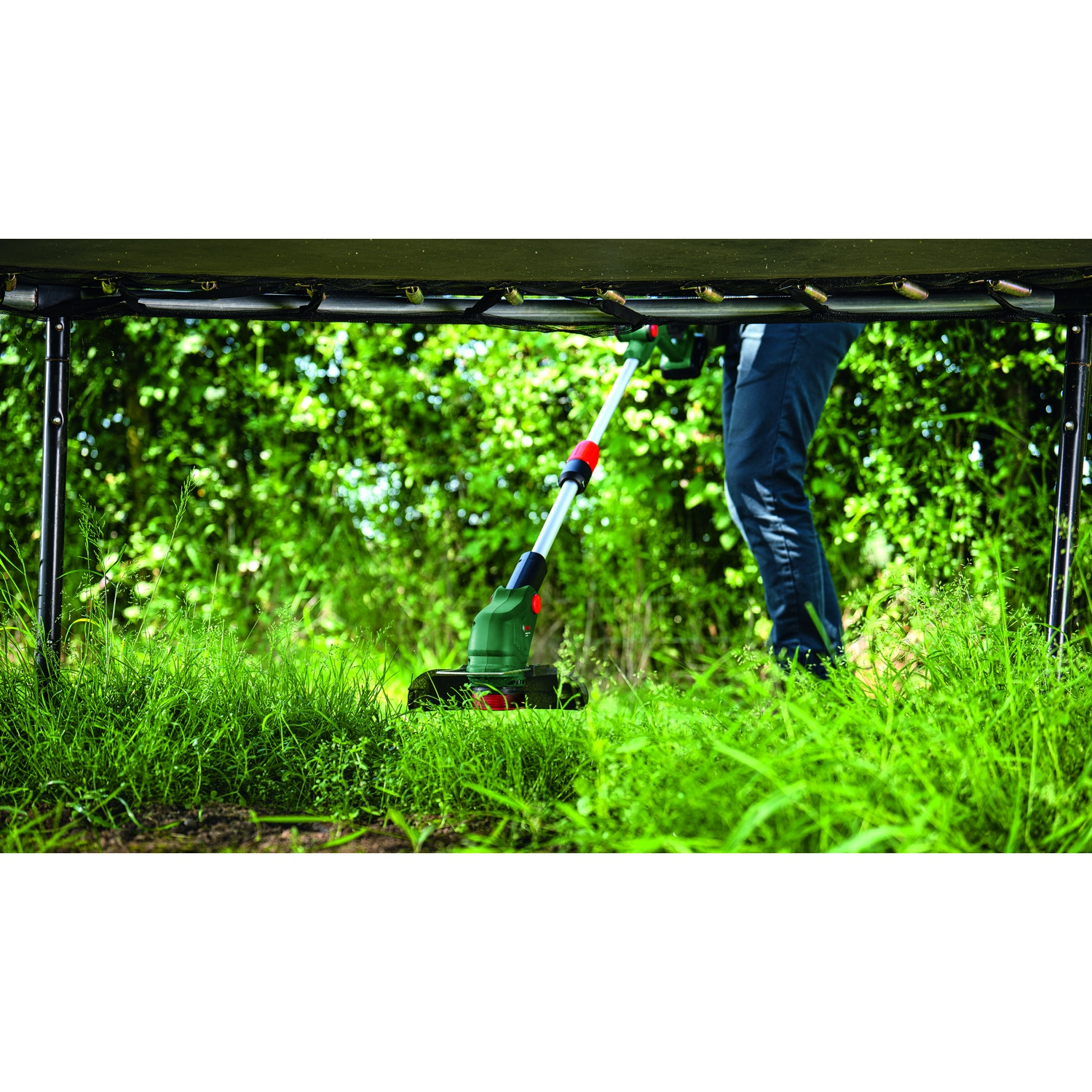 Una persona taglia l'erba con un decespugliatore. La scena è sotto un trampolino. Erba verde e fogliame sullo sfondo.