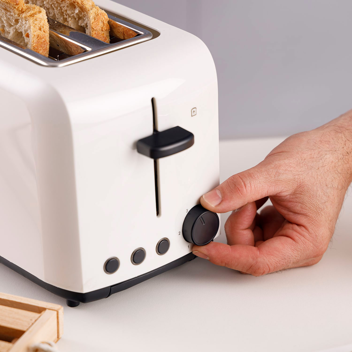 Una tostadora blanca con pan dentro. Una mano ajusta el dial de tostado. Una pequeña caja de madera en primer plano.