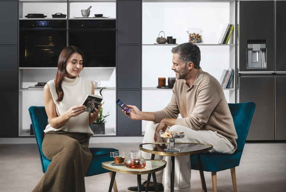 Férfi és nő modern konyhában. A nő könyvet olvas, a férfi telefont tart. Tea és sütemények az asztalon.