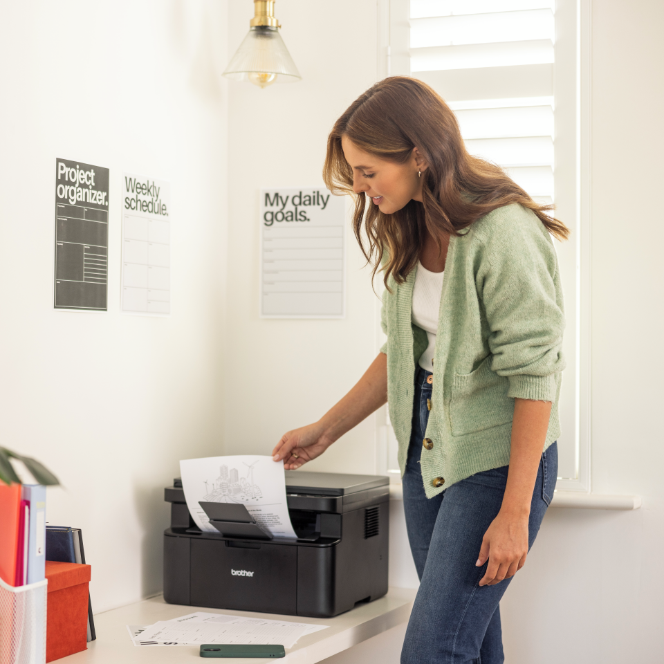 Kobieta w zielonym kardiganie wyjmuje wydrukowaną kartkę z czarnej drukarki. Drukarka stoi na białym biurku w pokoju.