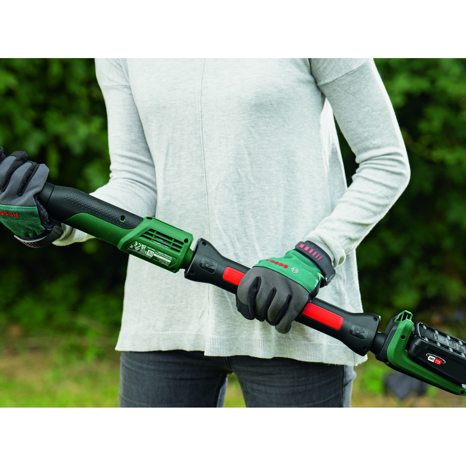 Una persona tiene in mano un attrezzo da giardinaggio verde e nero, indossando guanti. Lo sfondo è un fogliame verde sfocato. L'attrezzo ha un accento rosso.