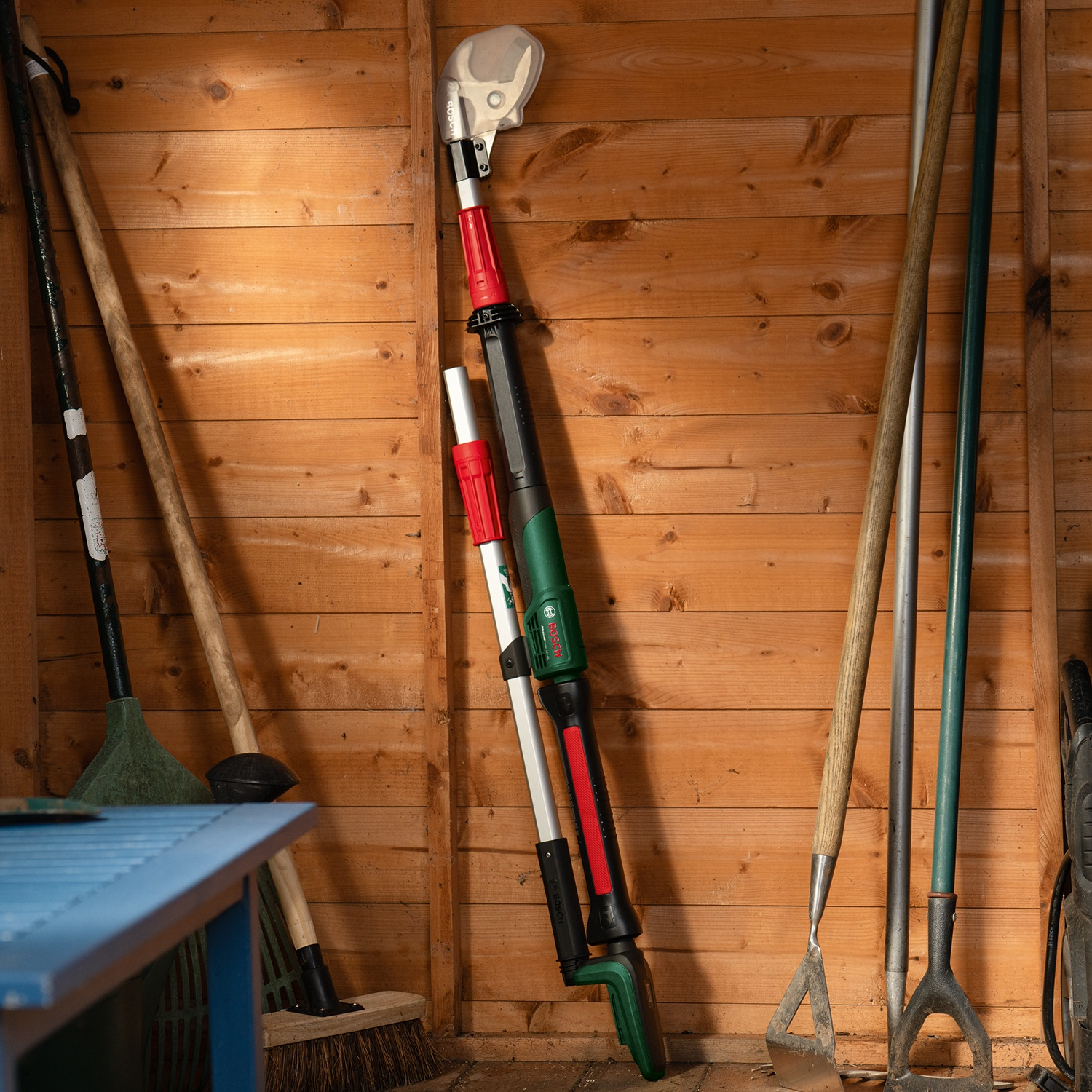 Interno di un capanno di legno con attrezzi da giardinaggio. Un elettroutensile è al centro, circondato da rastrelli, zappe e una scopa. Un tavolo blu è in primo piano.