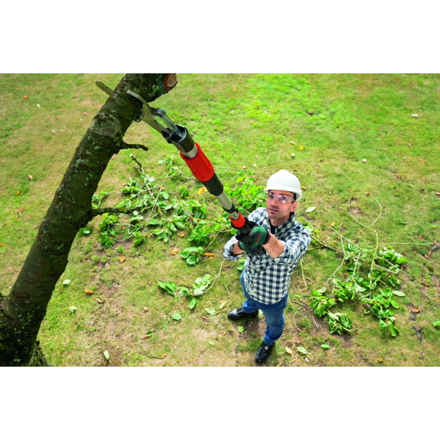 Un uomo con una camicia a quadri e un casco sta tagliando un ramo d'albero con un seghetto telescopico. Erba verde e rami sullo sfondo.
