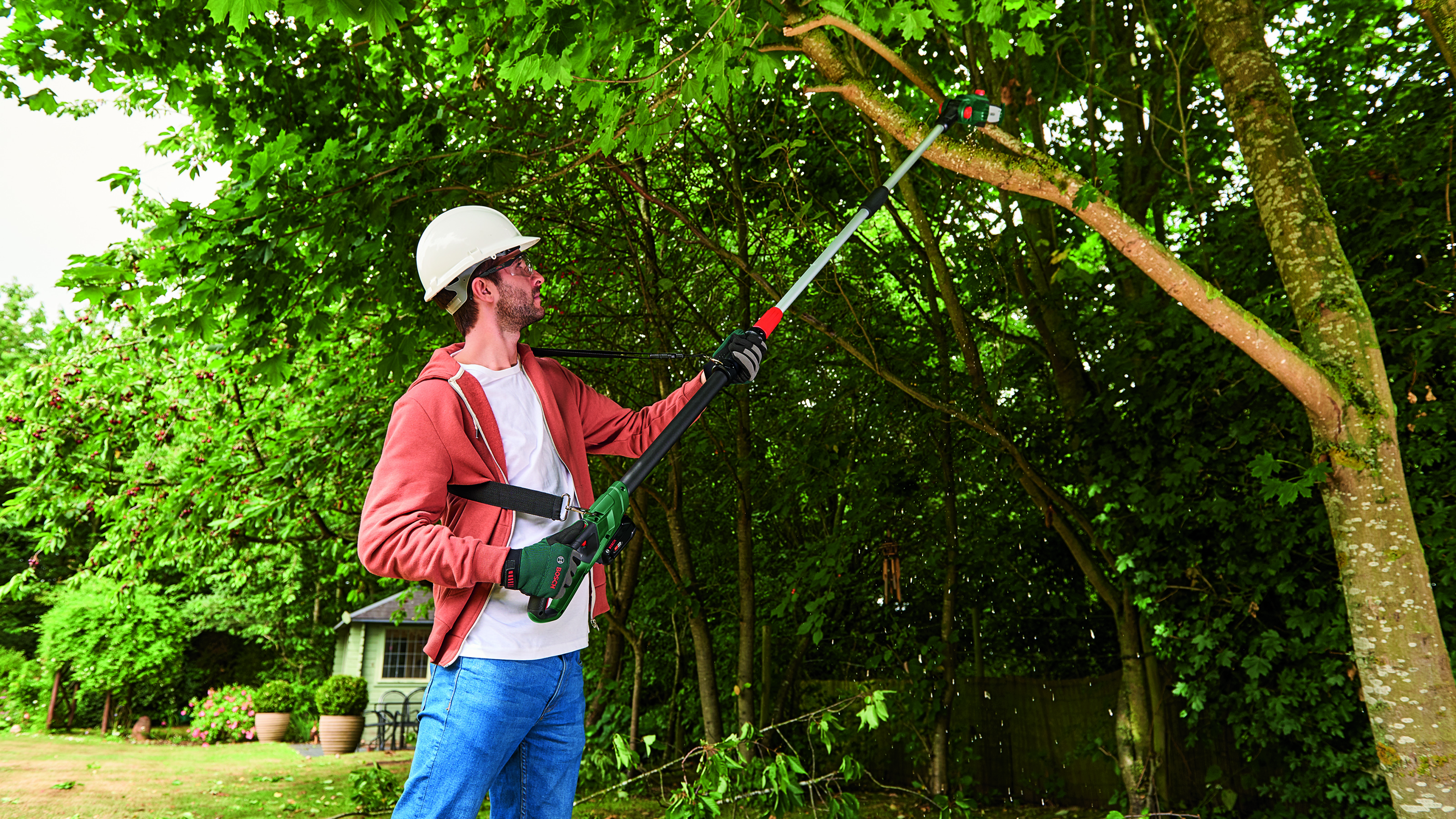 Un uomo con un casco di sicurezza usa un seghetto per tagliare un albero. Indossa una giacca e jeans. Lo sfondo include alberi e un edificio.