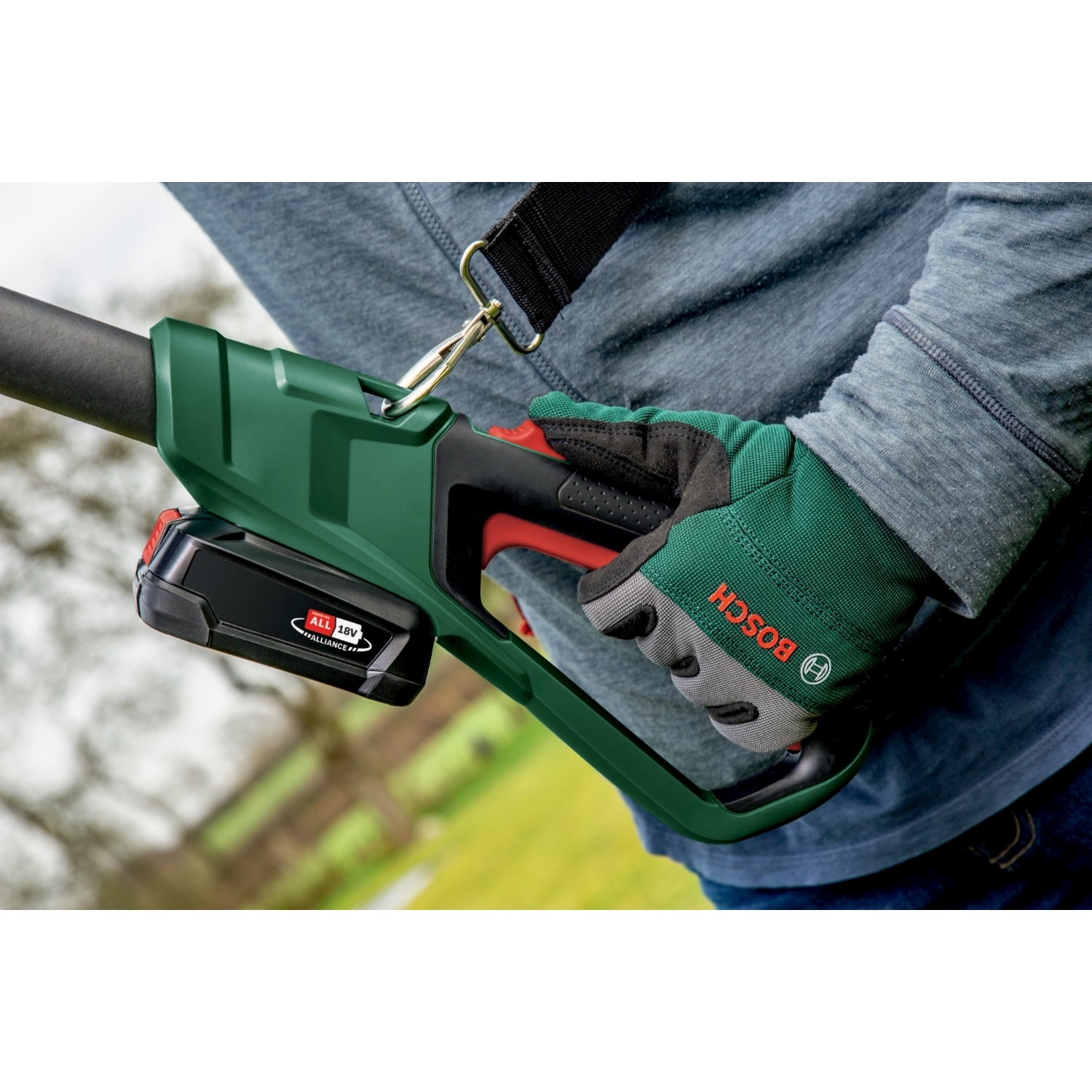 Una persona tiene in mano un attrezzo Bosch verde. La persona indossa una camicia grigia e guanti verdi. L'attrezzo ha una batteria e un cinturino neri.