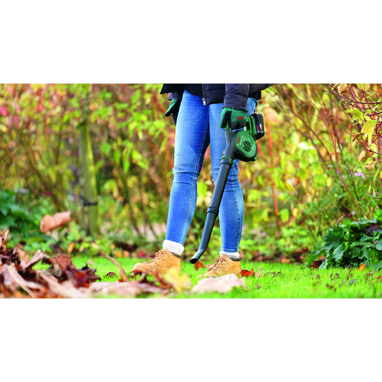 Una persona in jeans e stivali usa un soffiatore di foglie su un prato con foglie cadute. Fogliame verde sullo sfondo.