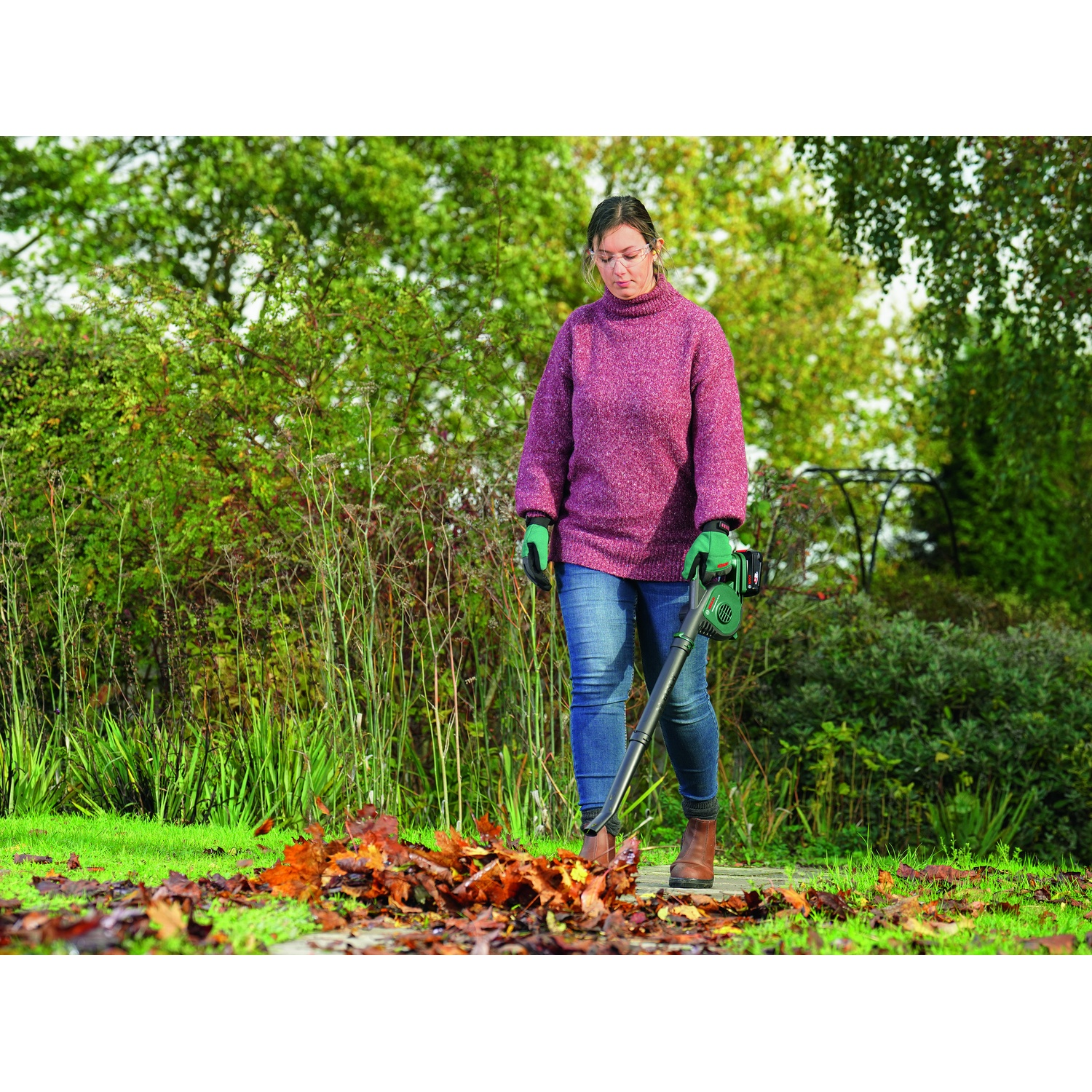 Una donna in maglione viola soffia le foglie con un soffiatore in giardino. Foglie marroni a terra, alberi verdi sullo sfondo.
