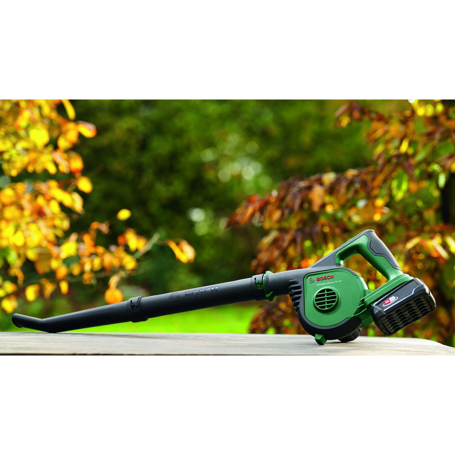 Un soffiatore per foglie Bosch verde e nero è appoggiato su una superficie di legno, con sfondo sfocato di fogliame.