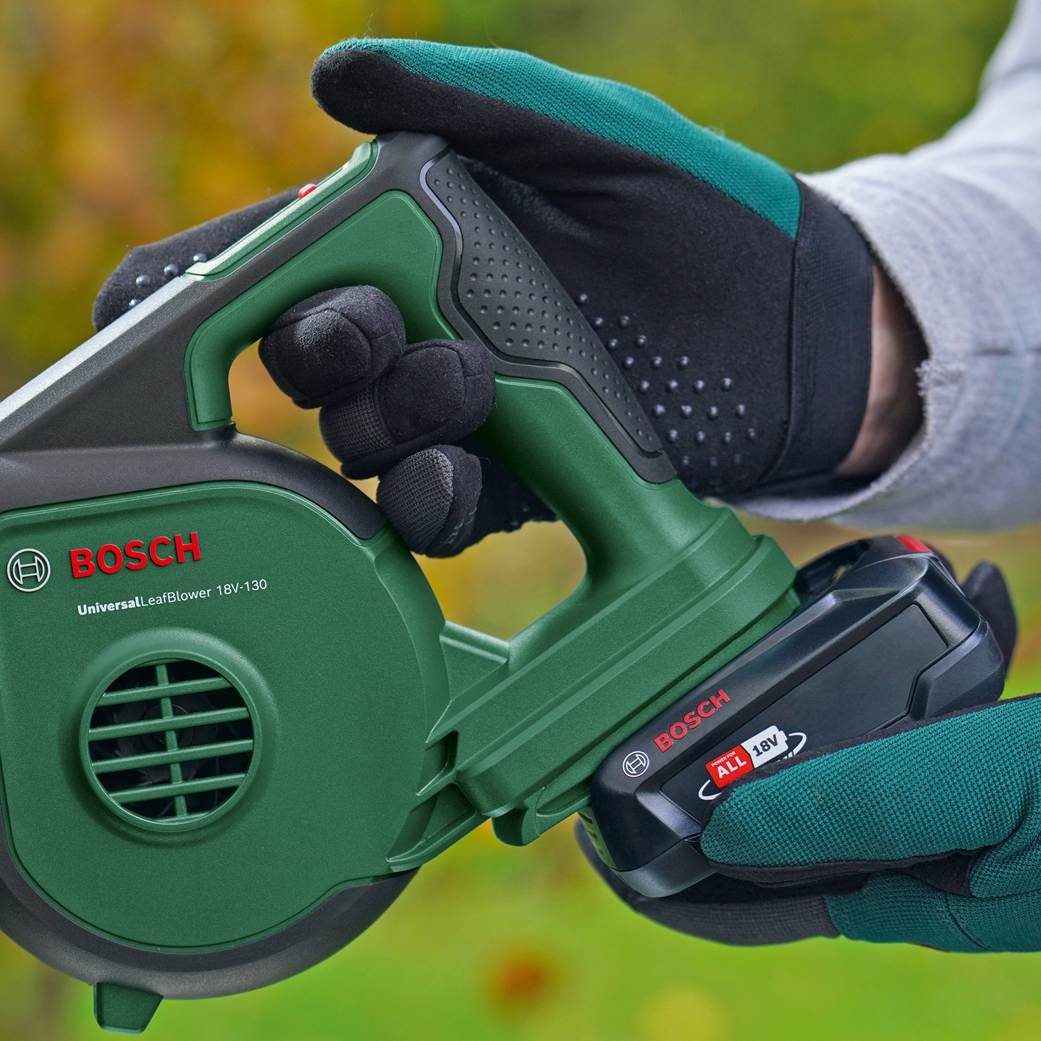 Una persona tiene in mano un soffiatore di foglie Bosch verde con batteria. La persona indossa guanti da lavoro. Lo sfondo è un fogliame sfocato.