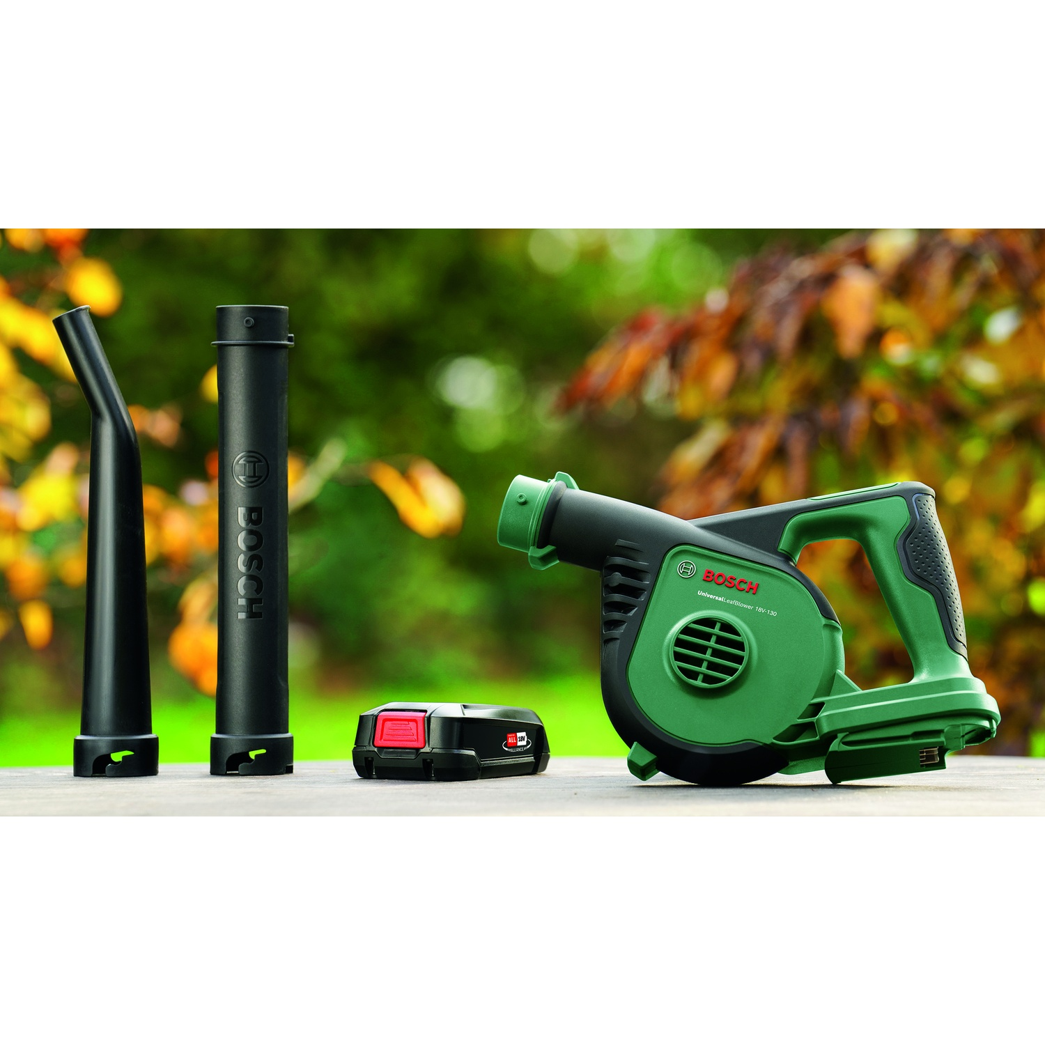 Un soffiatore per foglie Bosch verde e nero con accessori e batteria, su sfondo sfocato verde e marrone.