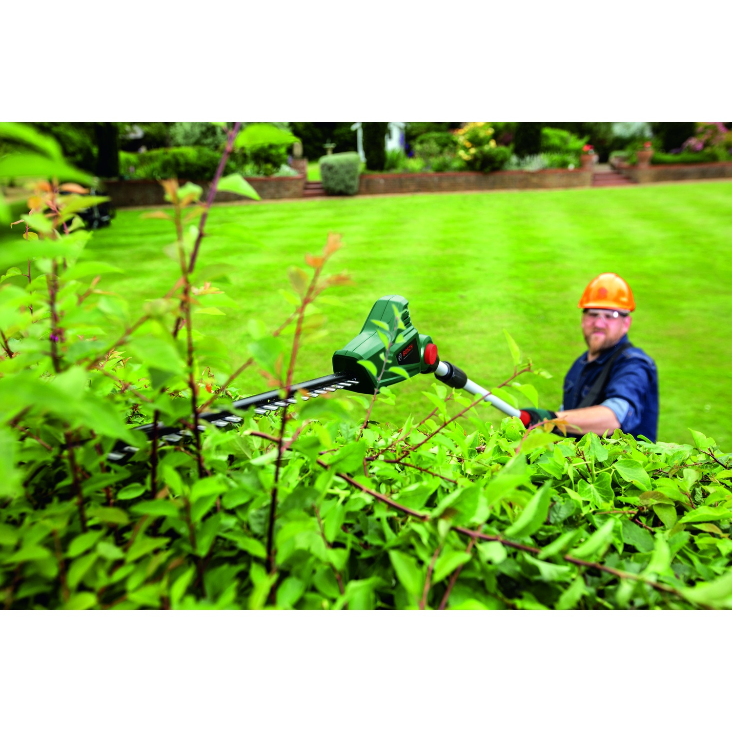 Un uomo in giardino, con un casco, taglia una siepe con un tagliasiepi elettrico. Erba verde e altre piante sullo sfondo.