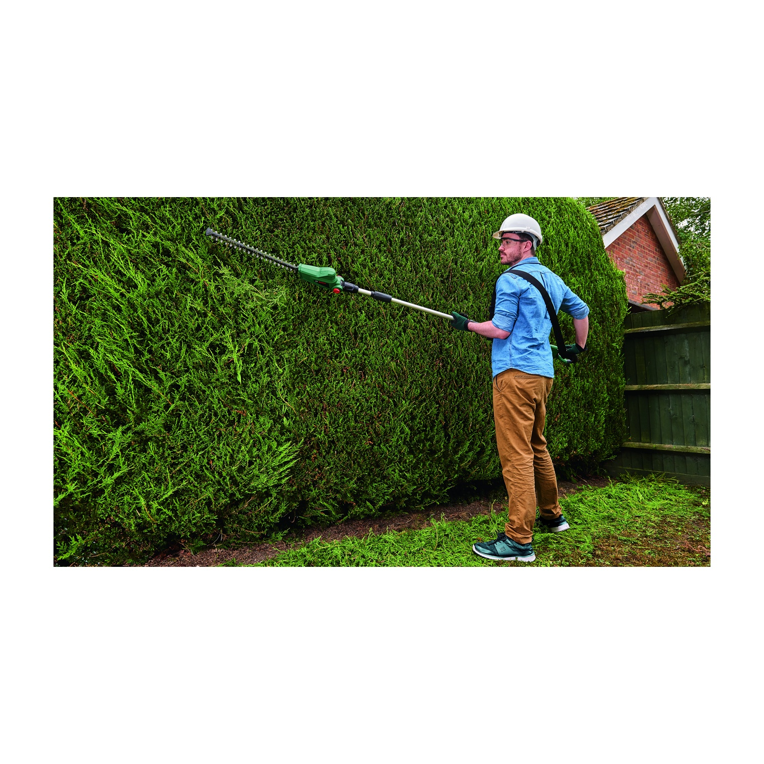 Un uomo pota un'alta siepe con un tagliasiepi elettrico. Indossa un elmetto, una camicia blu e pantaloni marroni, in giardino.